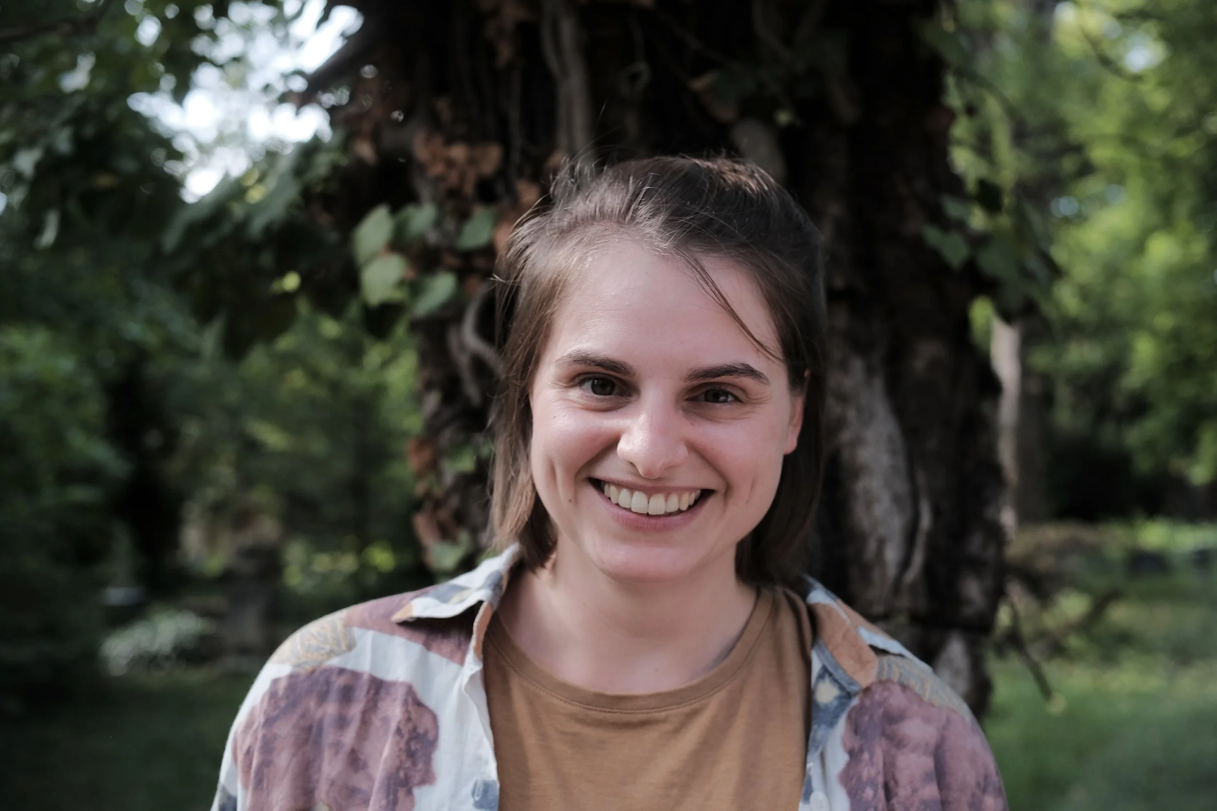 Smiling young woman in a plaid shirt standing outdoors in front of a tree with green leaves.