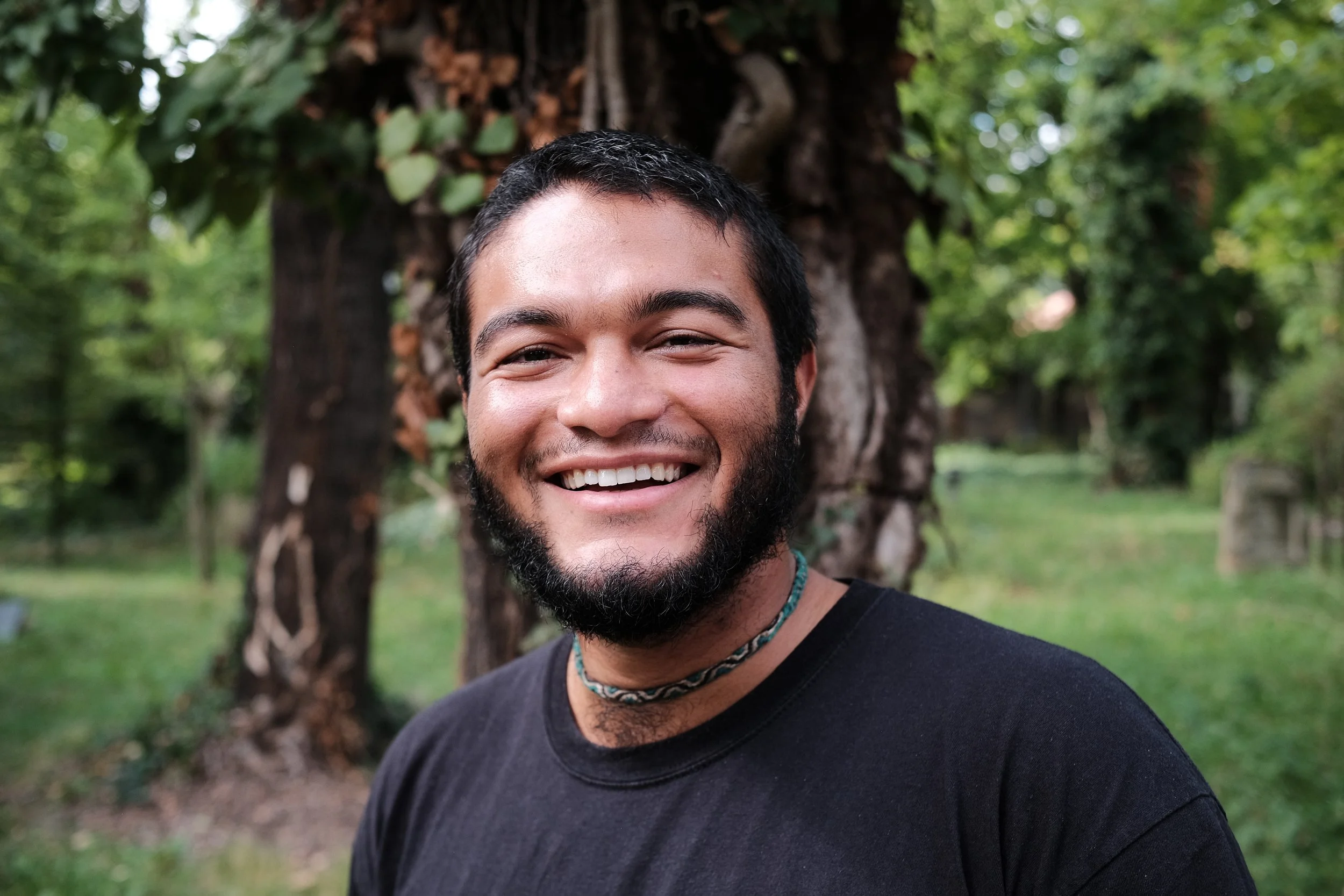 A young man with black hair and a beard smiling outdoors in front of a tree with green leaves.