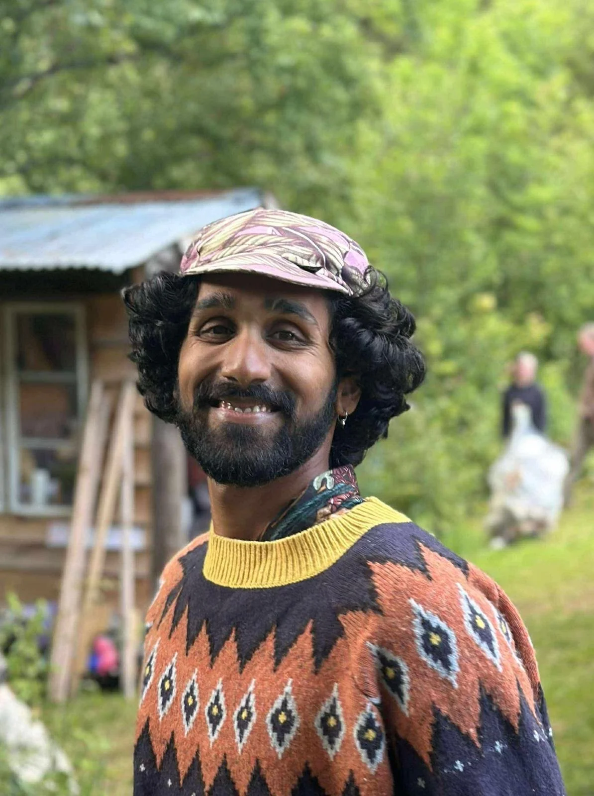 A man with curly black hair, a beard, and a small earring smiling outdoors. He is wearing a colorful sweater with a geometric pattern, a yellow collar, and a patterned cap. There are trees and a blurred person in the background.