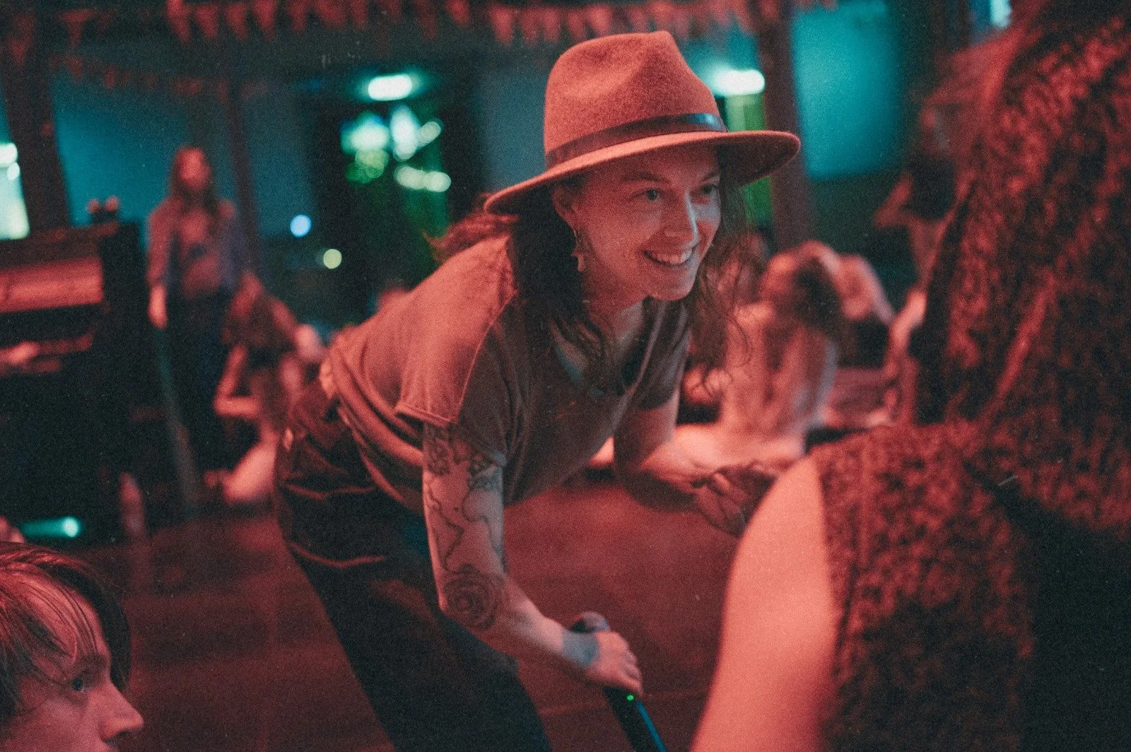 A woman with tattoos on her arm wearing a tan hat, smiling and leaning forward at a social gathering or party in a dimly lit venue with colorful lights.