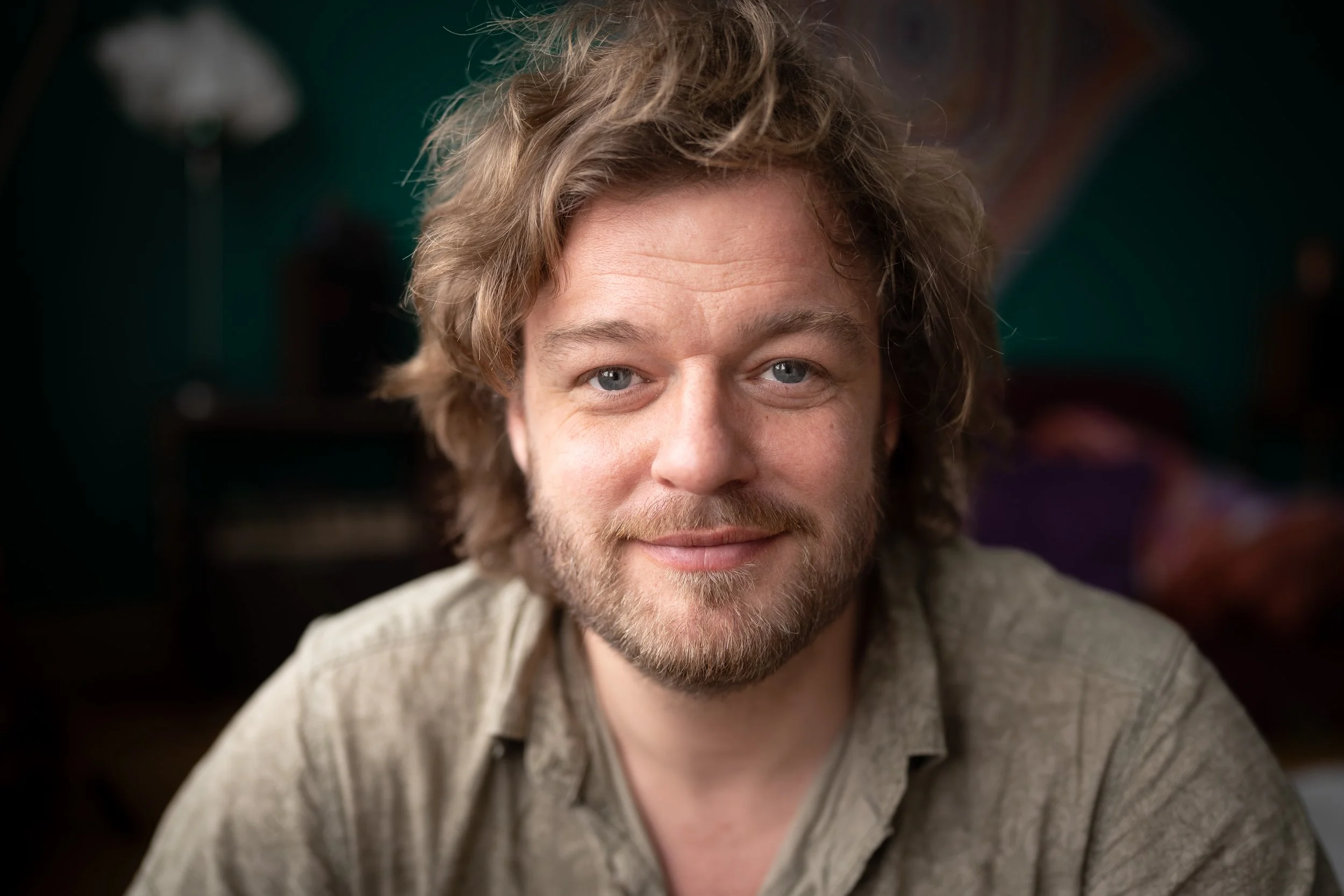 A close-up photo of a young man with wavy, blond hair and blue eyes, smiling slightly. He has a beard and is wearing a light-colored shirt with a textured pattern, set against a blurred dark background.