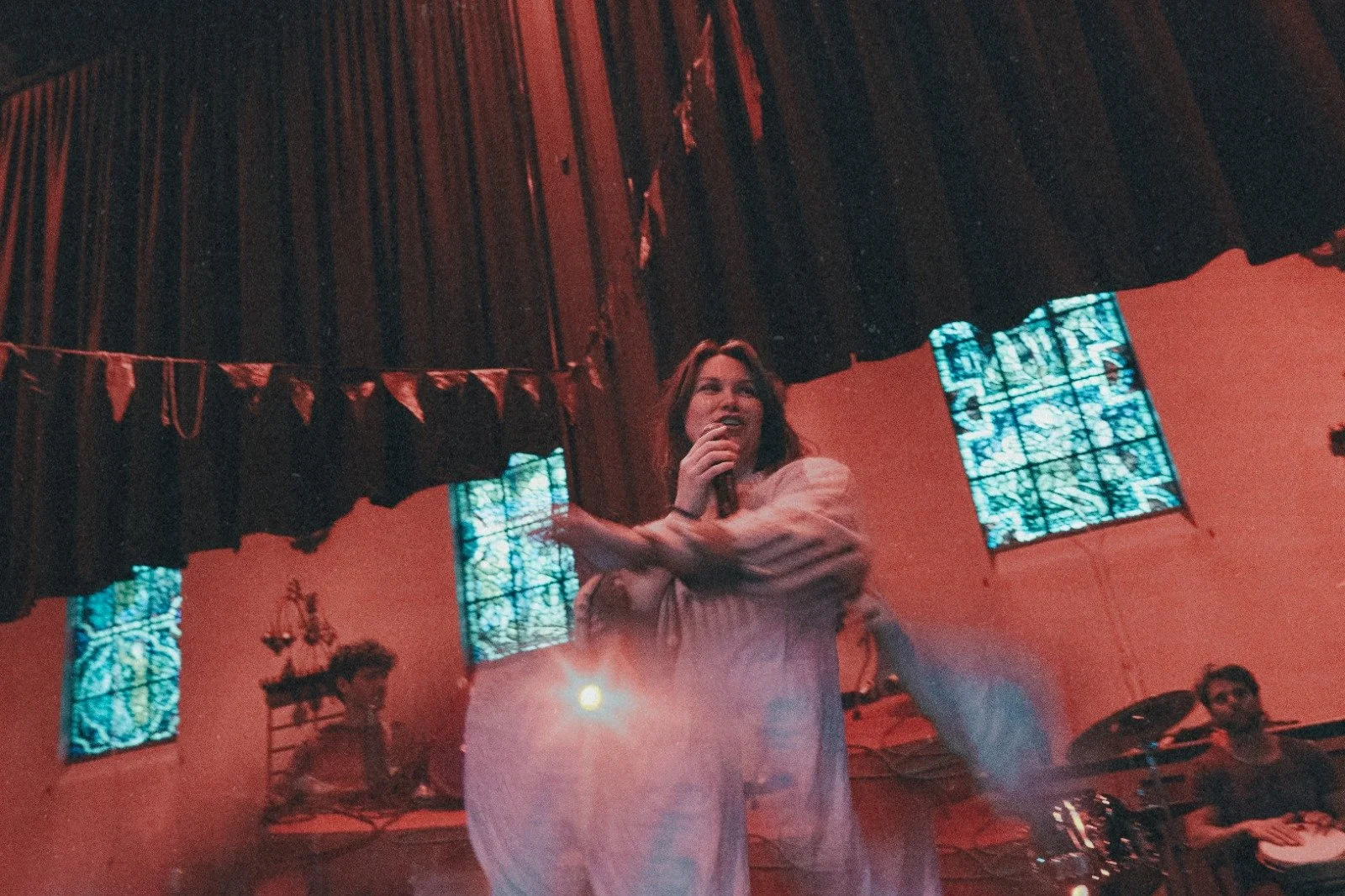 A woman with long brown hair is holding a microphone and talking or singing on stage. She is wearing a light-colored, long-sleeved shirt. There are stained glass windows behind her with blue and green patterns, and a musician playing drums can be seen to her right.