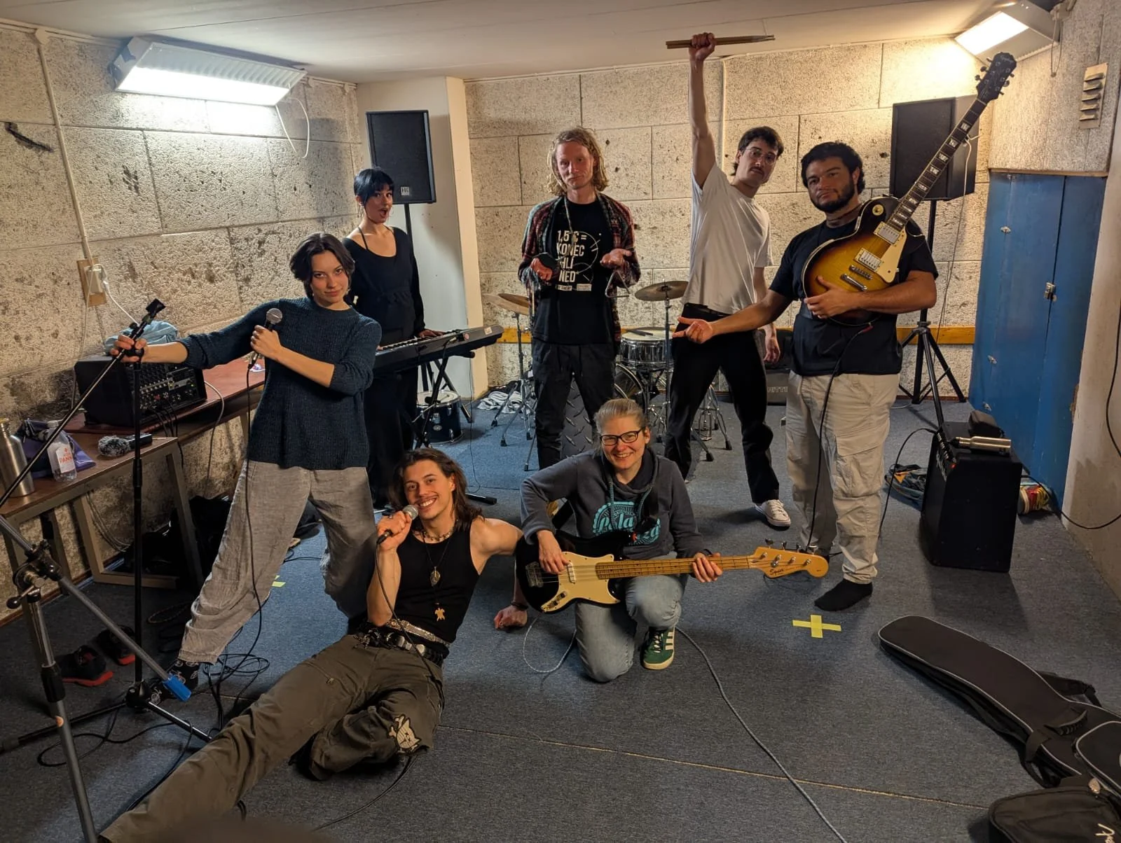 A group of seven young musicians preparing for a performance in a rehearsal room. They are holding musical instruments including guitars, a bass, and a keyboard, with some smiling and others making playful expressions.