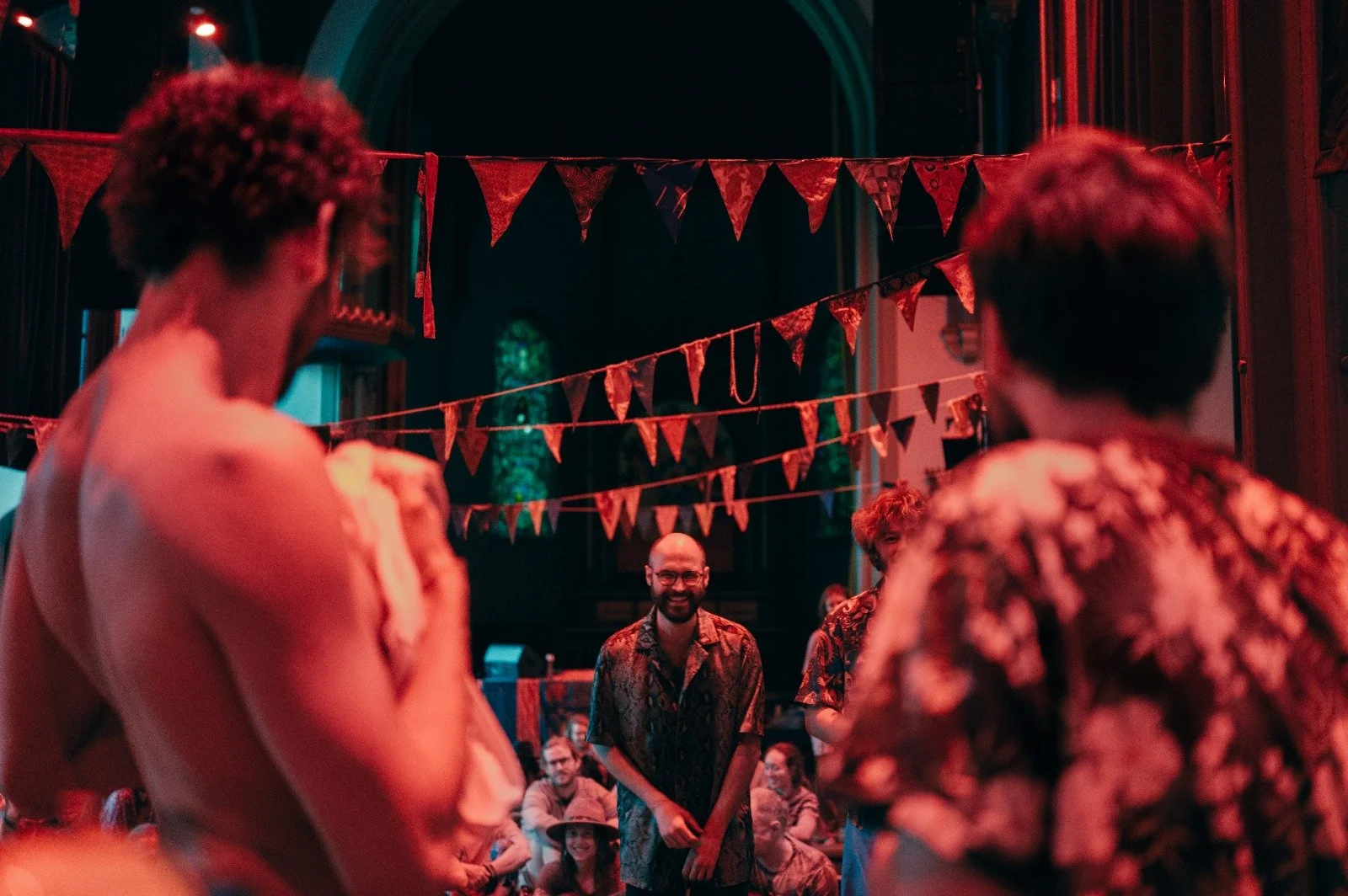People socializing at a party with red and orange lighting, red triangular banners hanging from the ceiling, in a large room with stained glass windows.