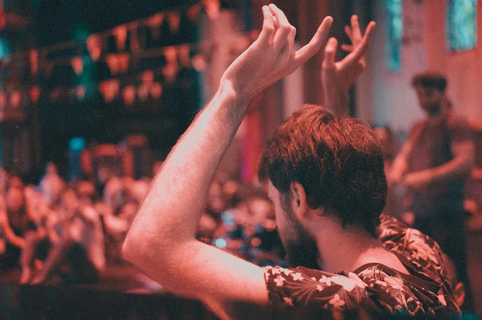 A person with short brown hair dancing at a crowded nightclub, raising their hand in the air, with colorful lights and other clubgoers in the background.
