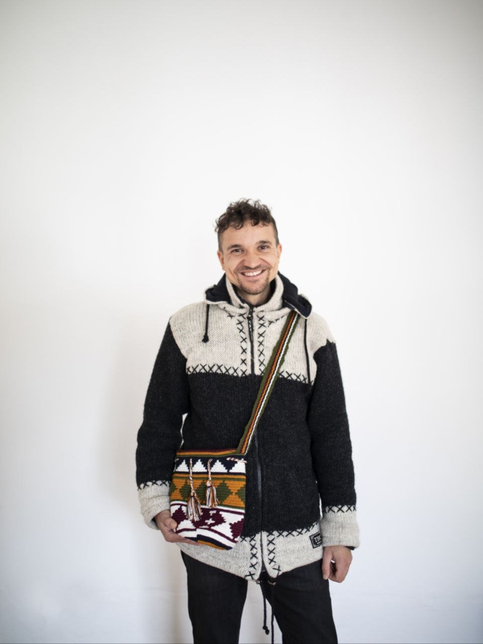 A man with short, curly brown hair and a beard stands smiling against a plain white wall. He is wearing a black and gray patterned knitted hoodie and has a colorful woven shoulder bag with geometric patterns.