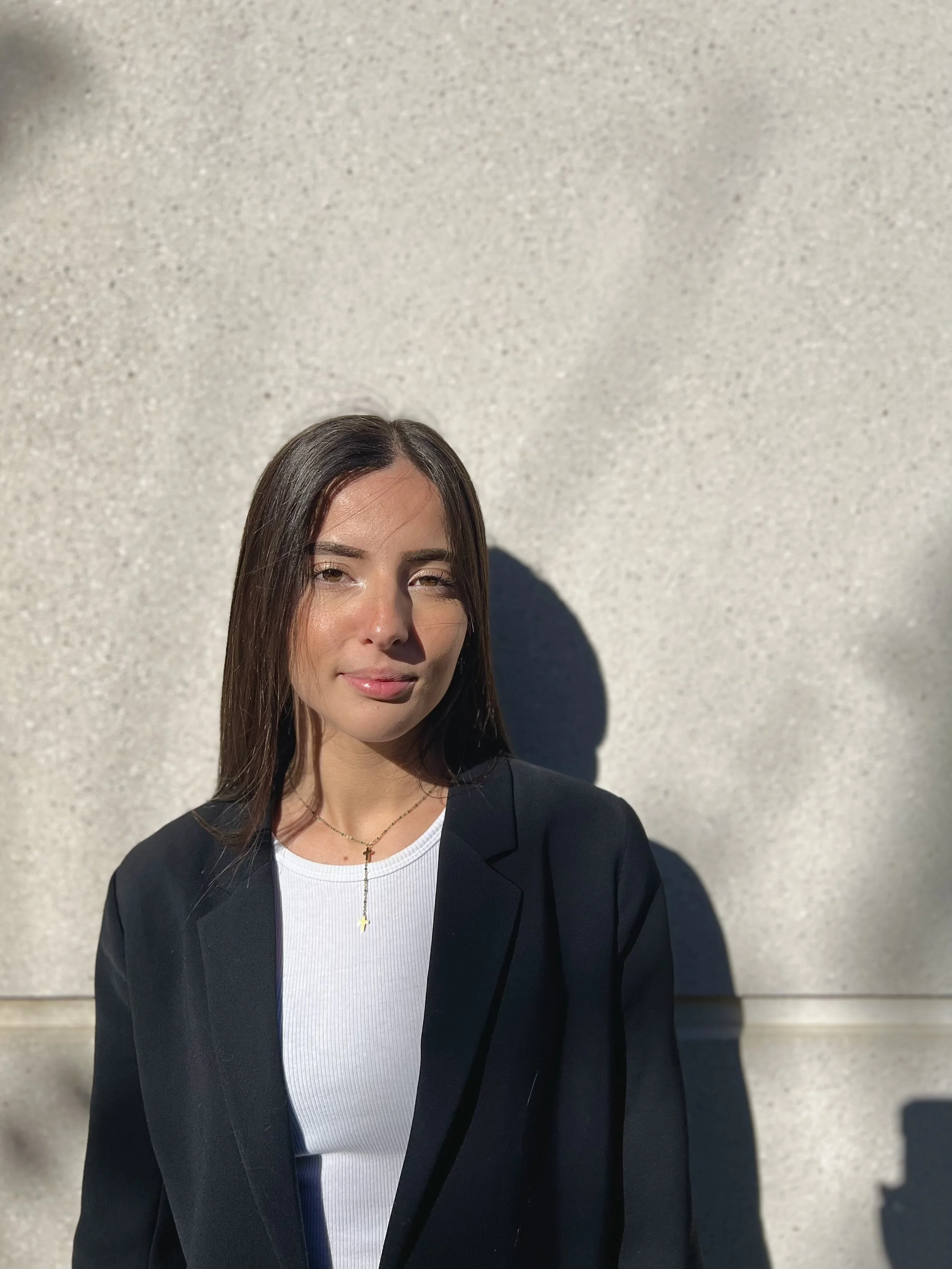 Jeune femme avec des cheveux bruns, portant un blazer noir et un t-shirt blanc, de portrait, en plein air, ensoleillé.