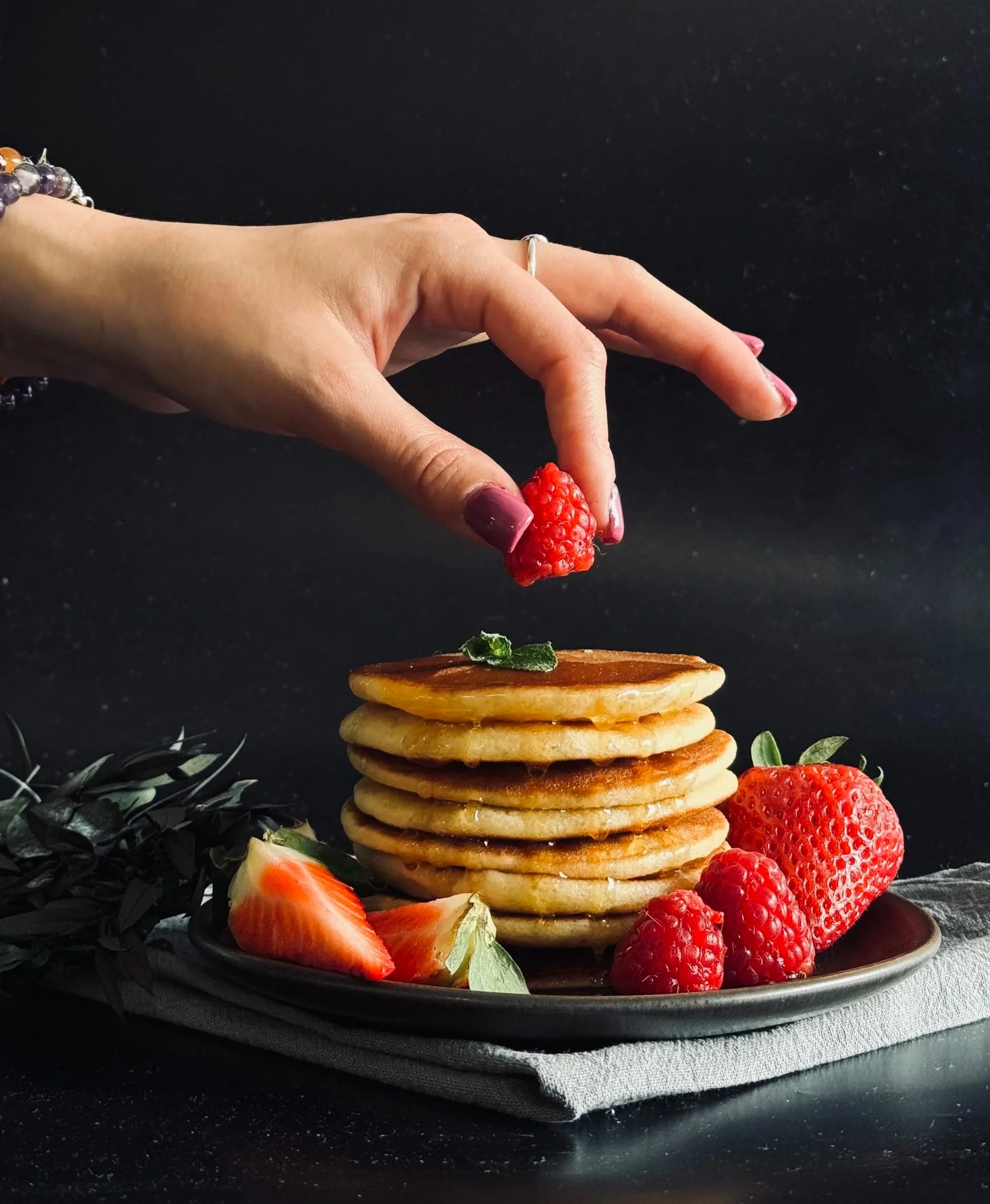 Une main tenant une framboise au-dessus d'un tas de pancakes avec des fraises autour sur une assiette noire.