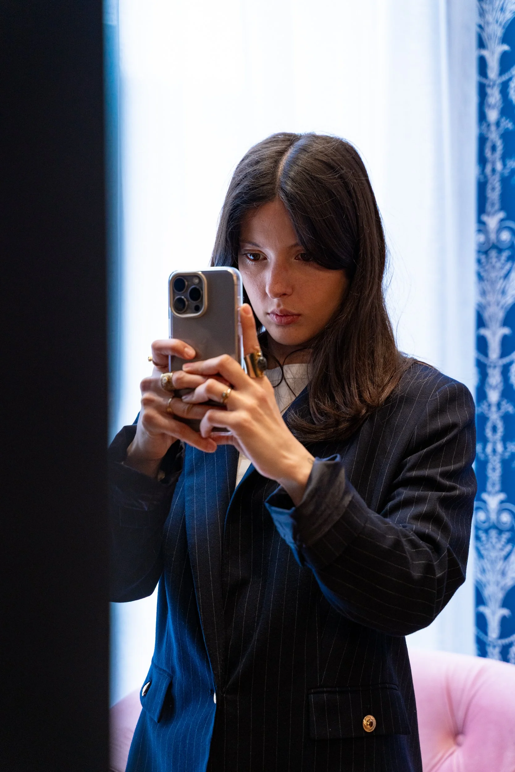 Une femme avec des cheveux bruns longs en costume noir, tenant un smartphone, prise en selfie dans un intérieur avec une lumière naturelle."}**assuming the goal is to describe the visual content for accessibility and SEO.**