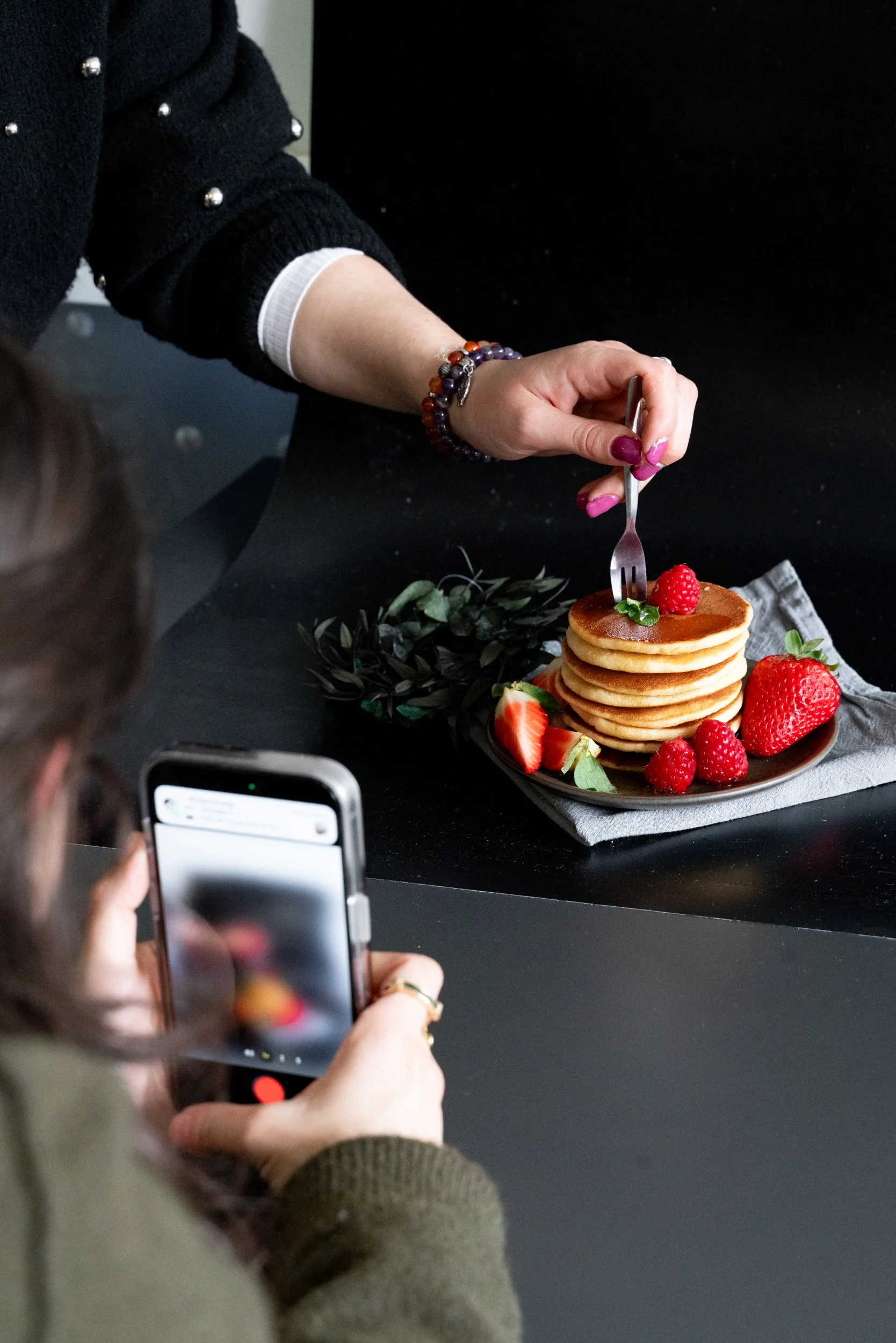 Une personne prend en photo un plateau de pancakes avec des fruits rouges, dont des fraises, des framboises et une mûre, sur une nappe grise. Une autre personne, dont seul le bras et la main sont visibles, tient une fourchette avec un pancake.