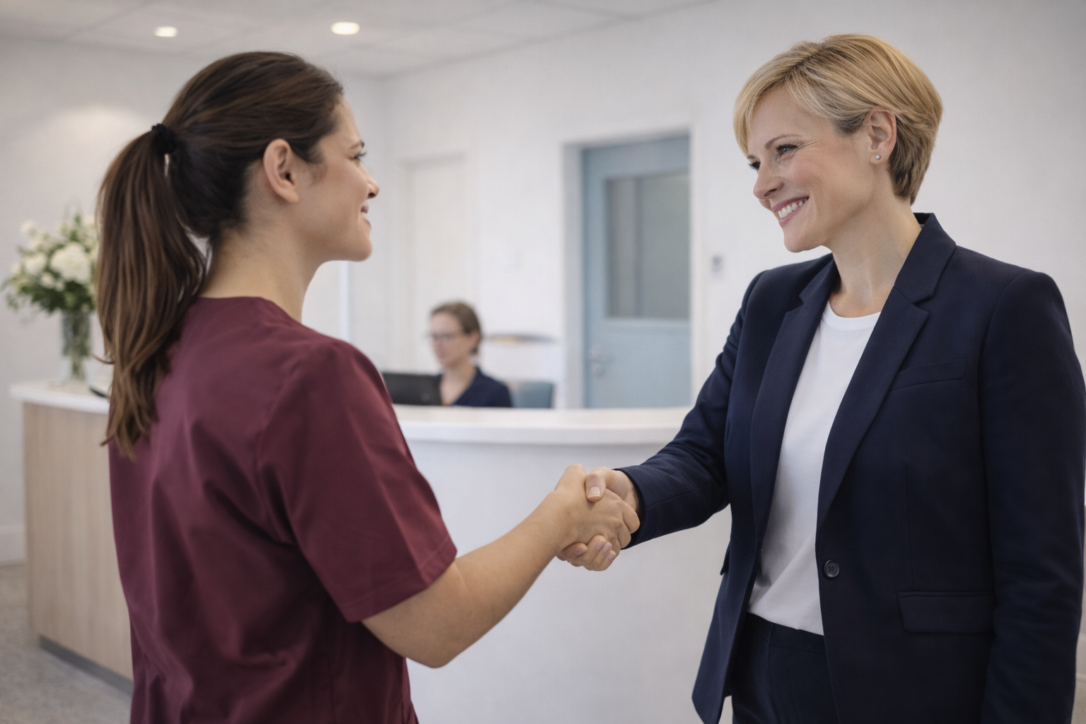 Two women shake hands in a professional setting, smiling at each other, with a receptionist or worker seated at a desk in the background.
