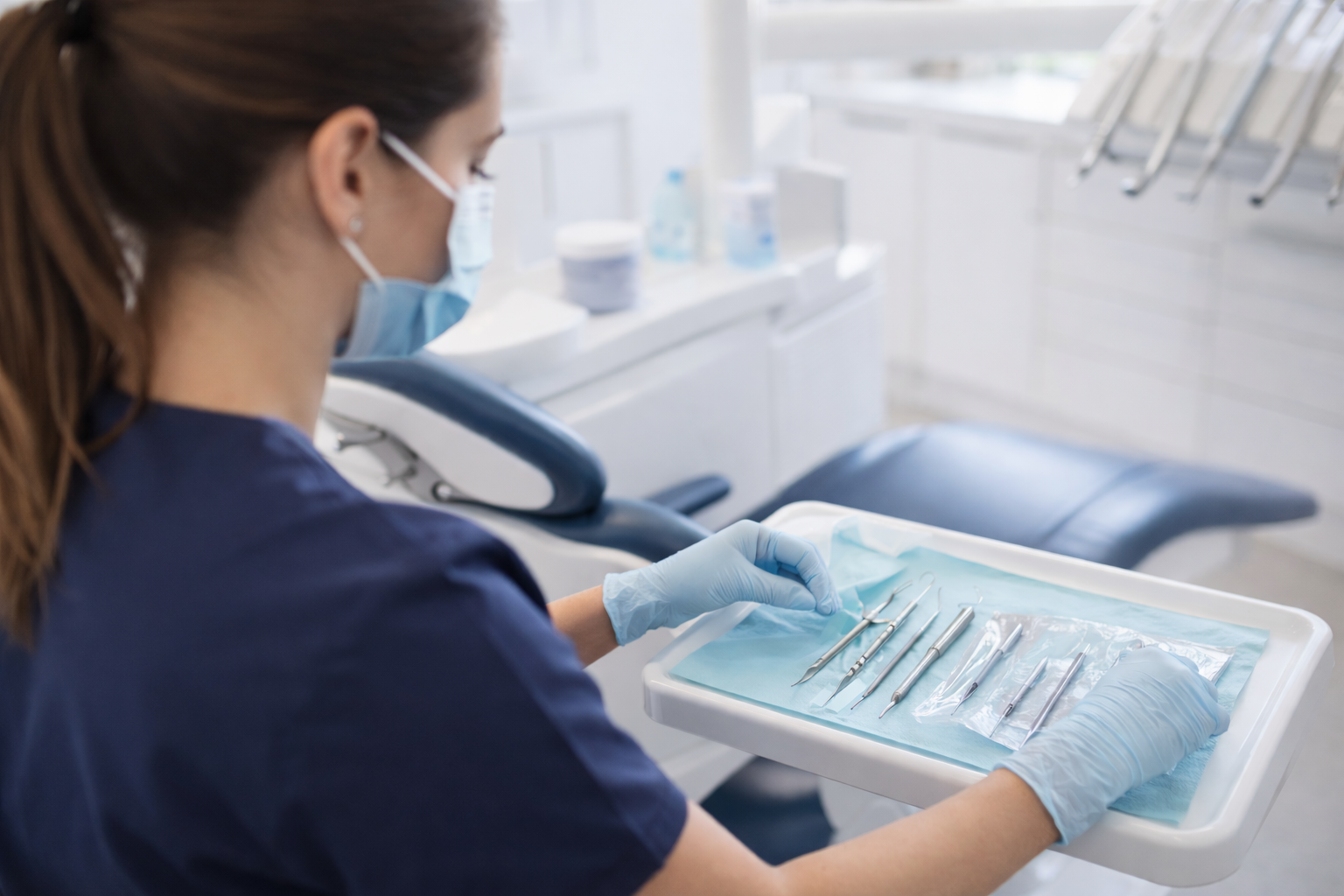 A female dental professional wearing a face mask and gloves handling dental tools on a tray in a dental clinic.
