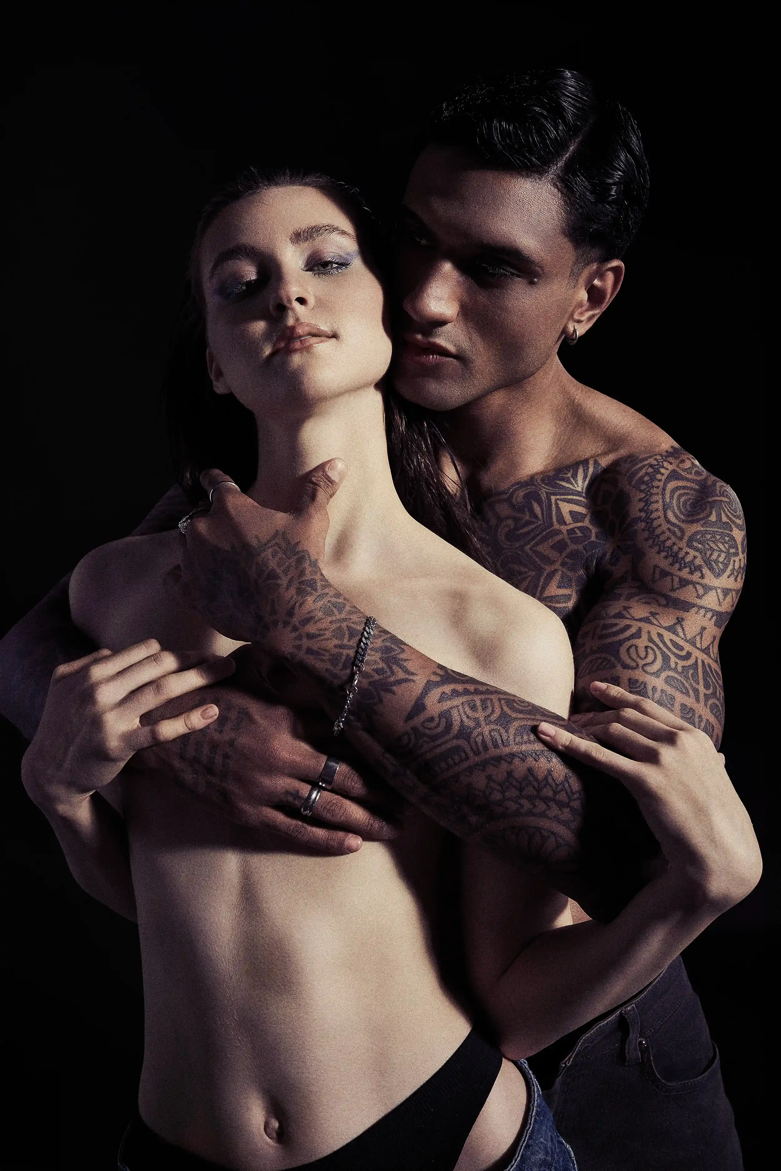 A man and woman embracing shirtless against a black background. The man has tattoos and is holding the woman, who has long dark hair and light skin, close to him.