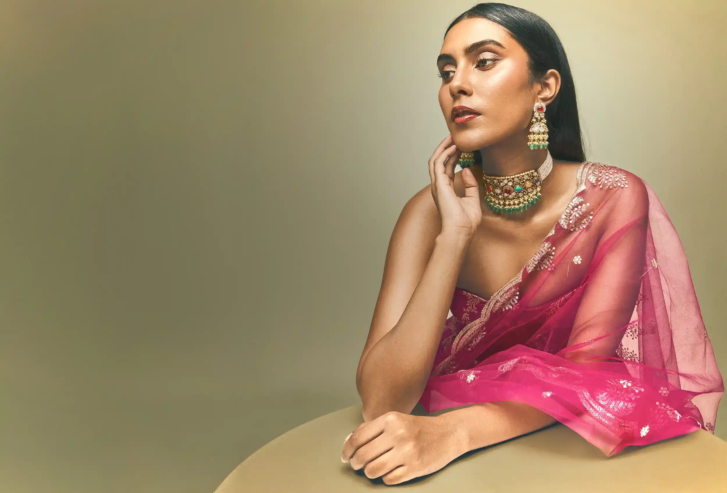 A woman dressed in traditional Indian attire with elaborate jewelry, sitting at a table with her hand resting on her chin, looking thoughtfully to the side. Anandclix, Bangalore photographer