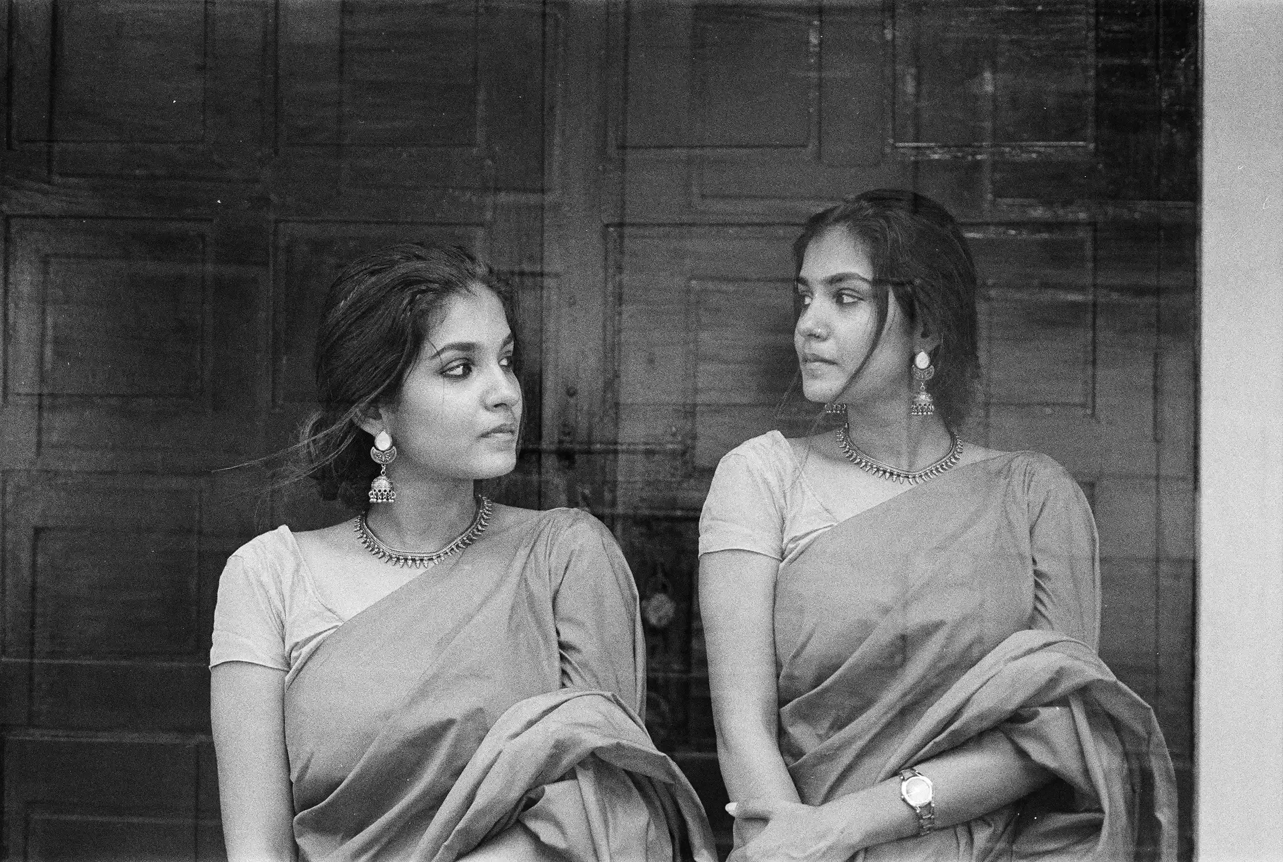 A black and white image of two women wearing sarees and jewelry, standing in front of a wooden paneled wall, looking at each other.