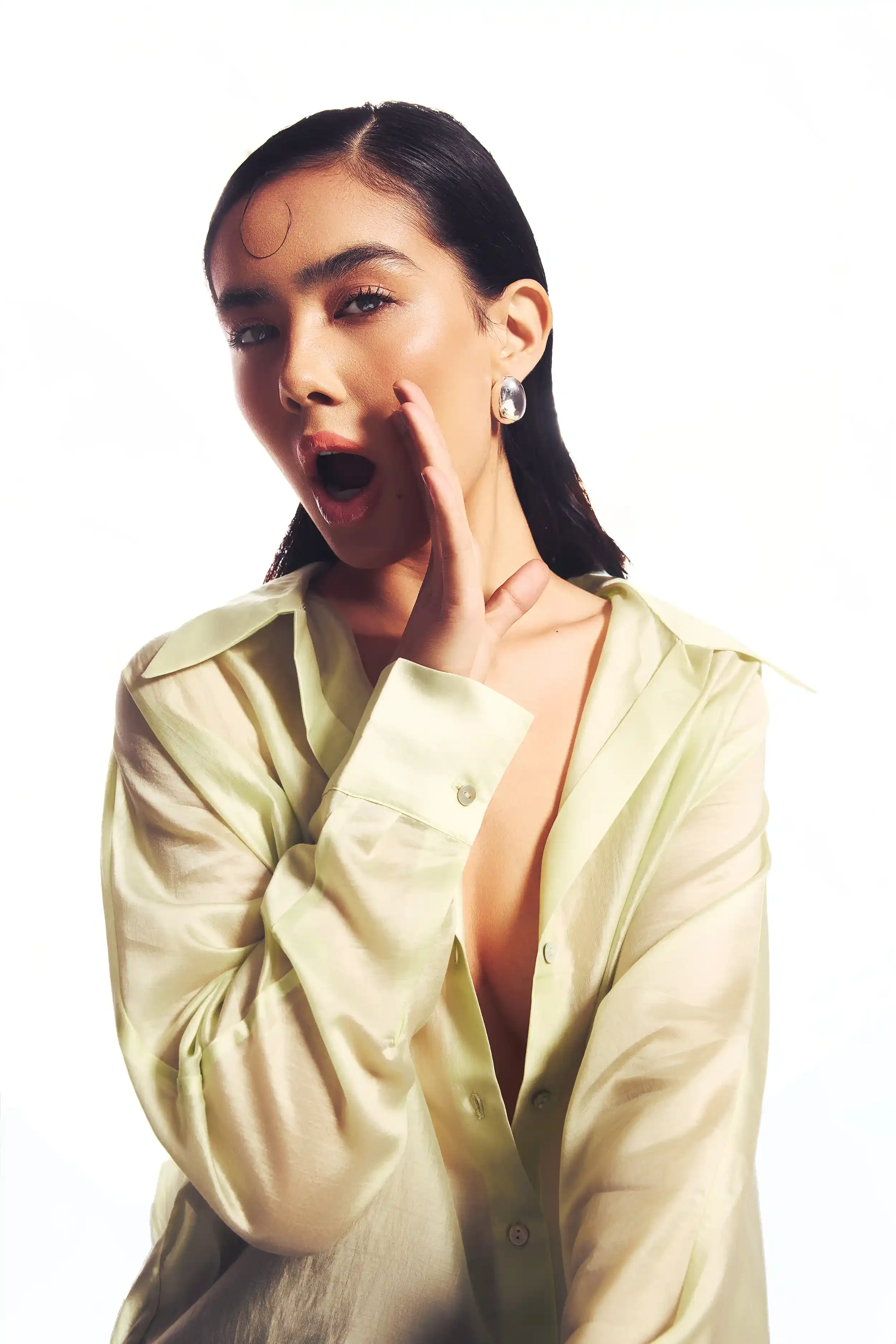 A woman with dark hair and fair skin wearing a light-colored satin shirt, looking surprised with her mouth open and one hand near her face, against a white background. Anandclix, Bangalore, advertising photographer, Fashion photographer, Editorial.