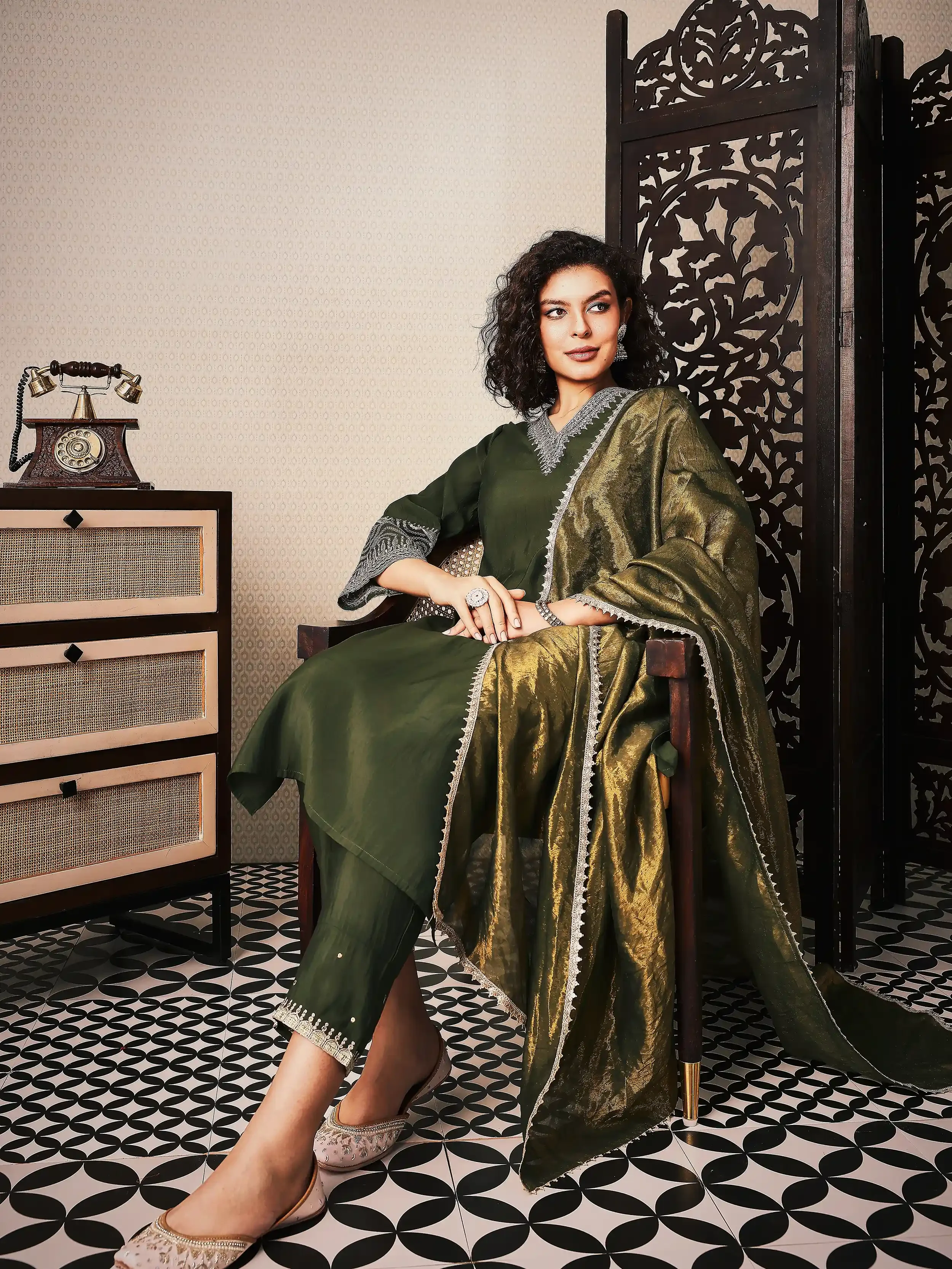 A woman wearing traditional Indian clothing, seated on a chair in a room with vintage decor, including a wooden screen and a cabinet with a rotary phone. Anandclix, Bangalore, advertising photographer, Fashion photographer, Editorial photographer.
