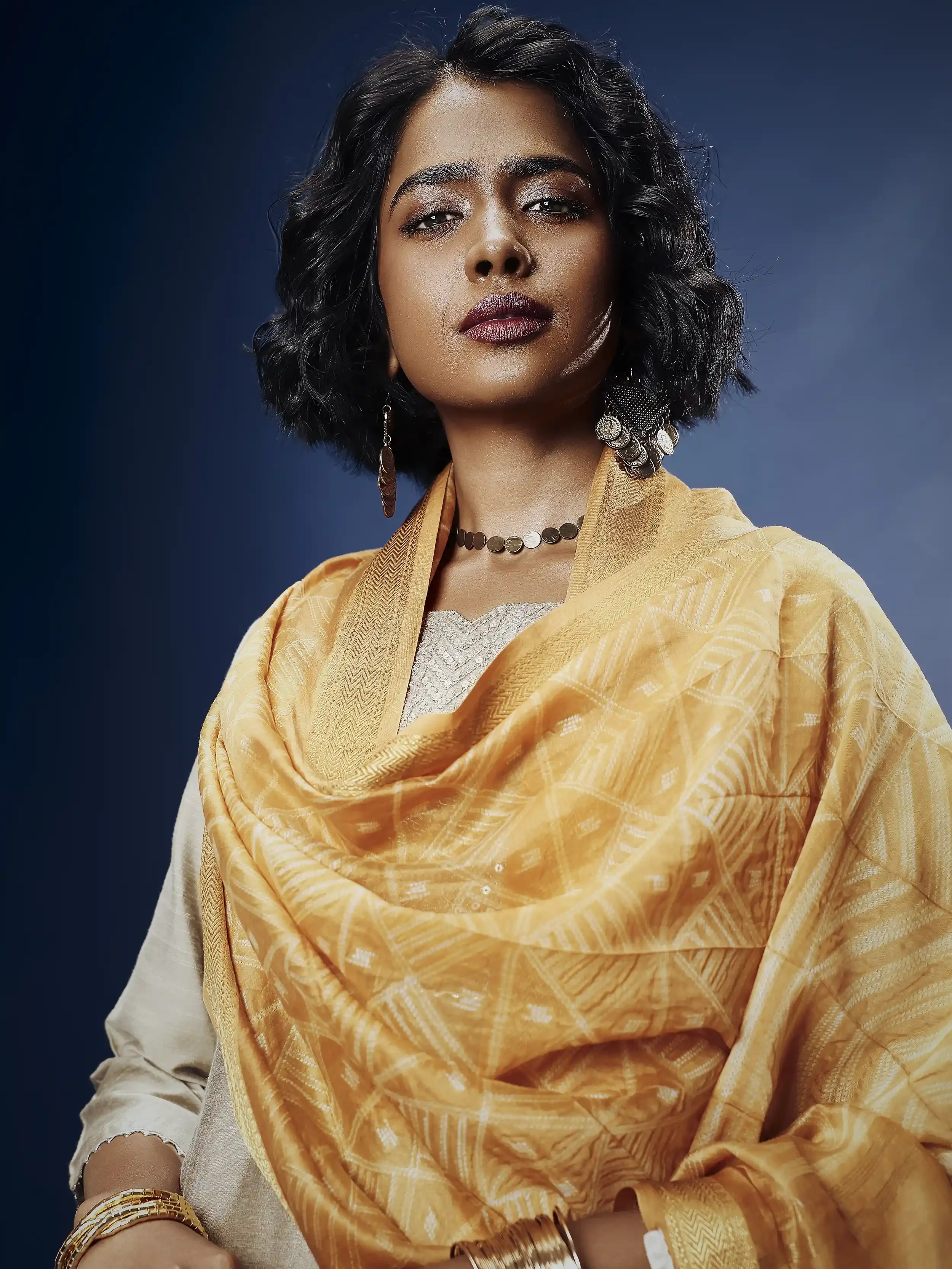 A woman with dark, wavy hair wearing a beige and orange traditional Indian saree with gold jewelry, posing against a dark blue background. Anandclix, Bangalore, advertising photographer, Fashion photographer, Editorial photographer.