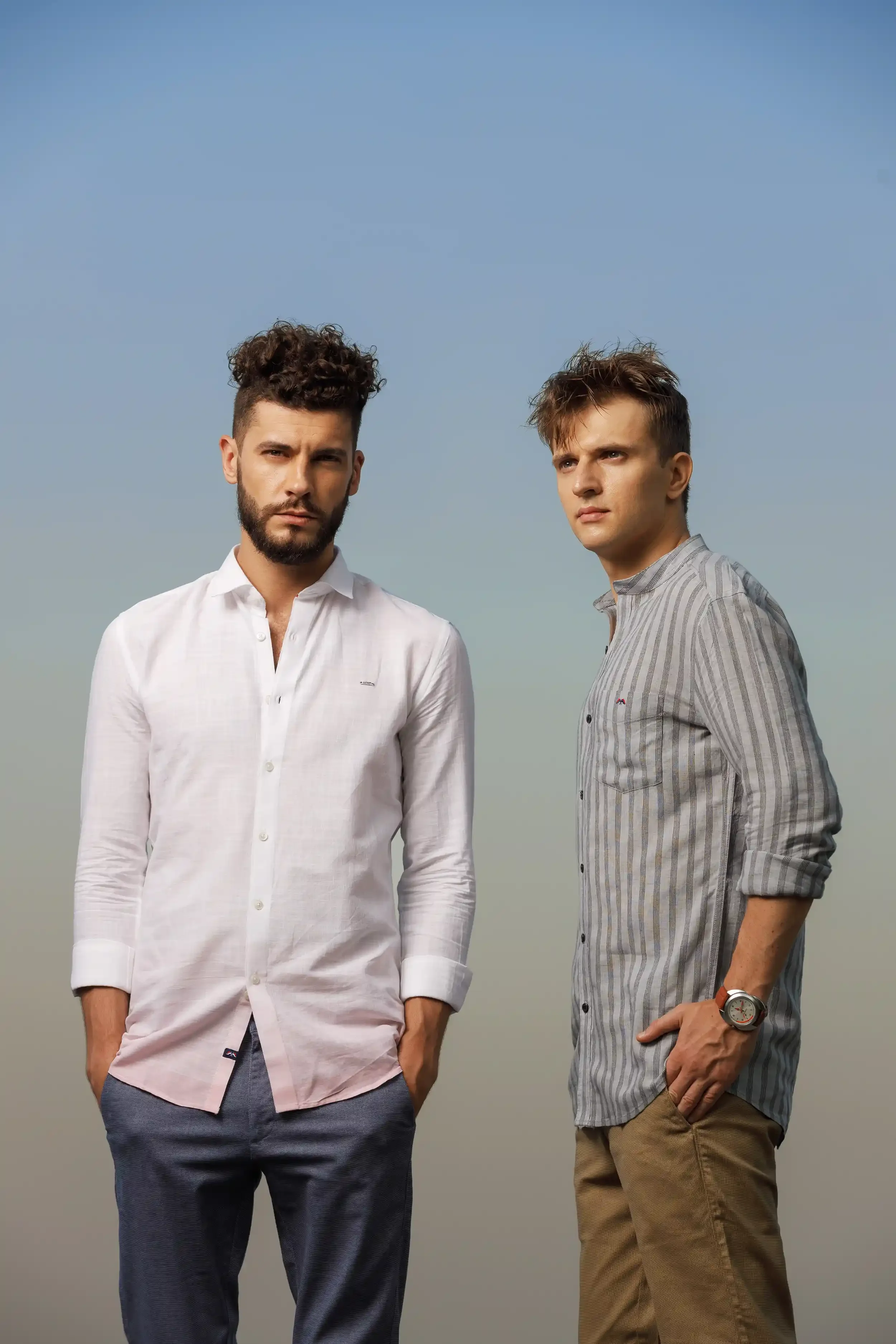 Two young men standing outdoors under a clear blue sky, looking serious. The man on the left has dark, curly hair and a beard, wearing a white button-up shirt and gray pants. The man on the right has tousled brown hair, wearing a gray striped shirt w