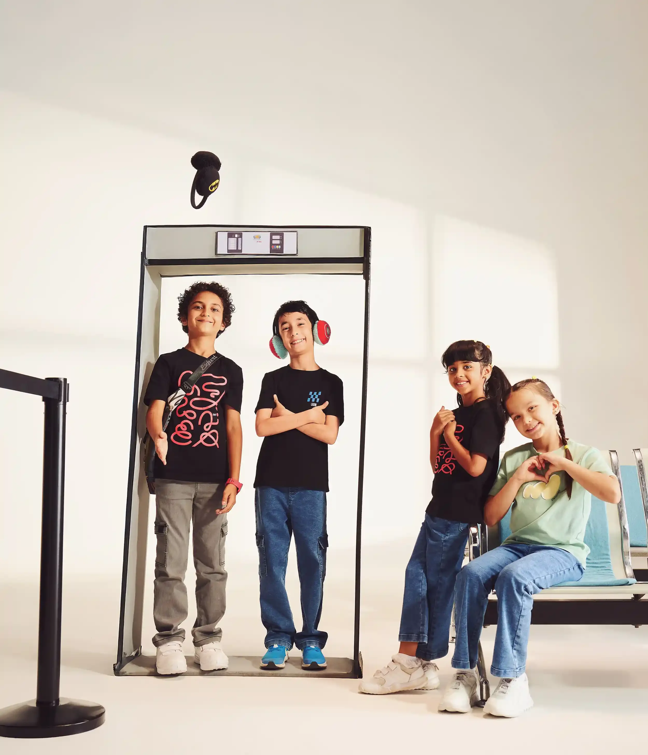 Four children standing and sitting in an airport security checkpoint, with one parent sitting on a bench. The children are smiling, some wearing headphones and making heart gestures with their hands. Anandclix, Bangalore, advertising photographer.