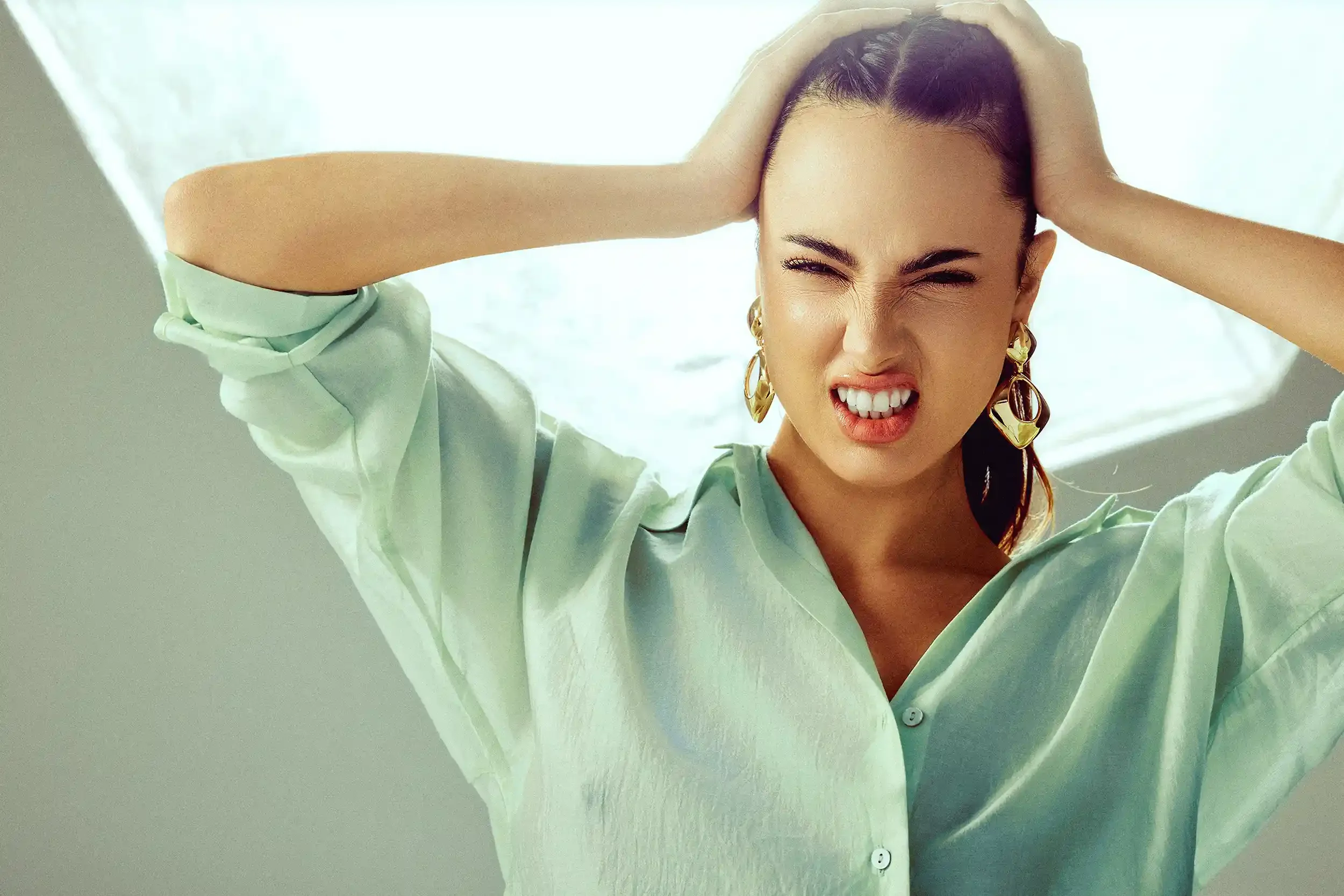 A woman with dark hair, wearing a light green satin shirt and large gold earrings, is holding her head with both hands and making a grimacing facial expression.