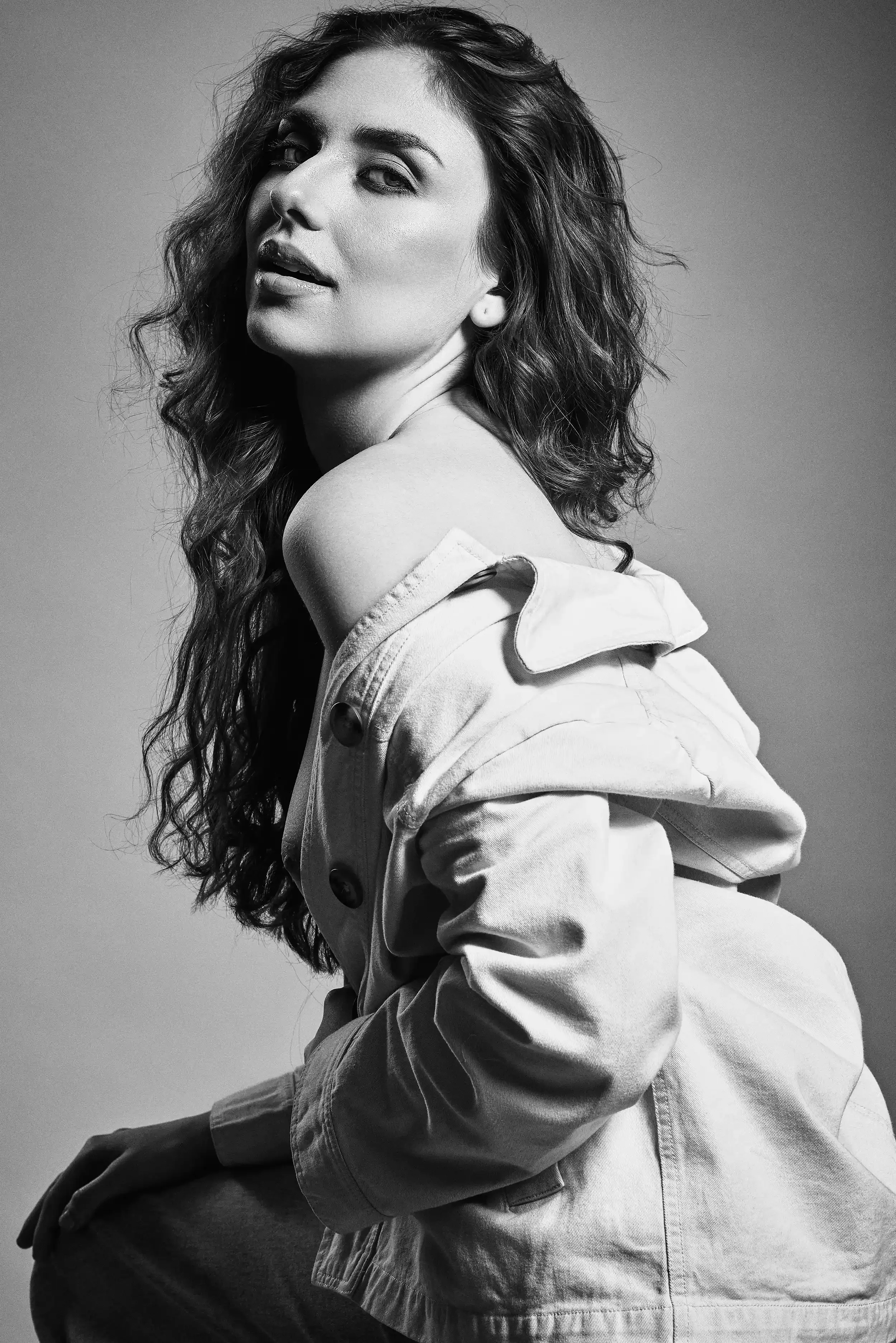 Black and white portrait of a woman with long, wavy hair, wearing an off-shoulder top and a denim jacket, posing with her head slightly tilted and a confident expression.