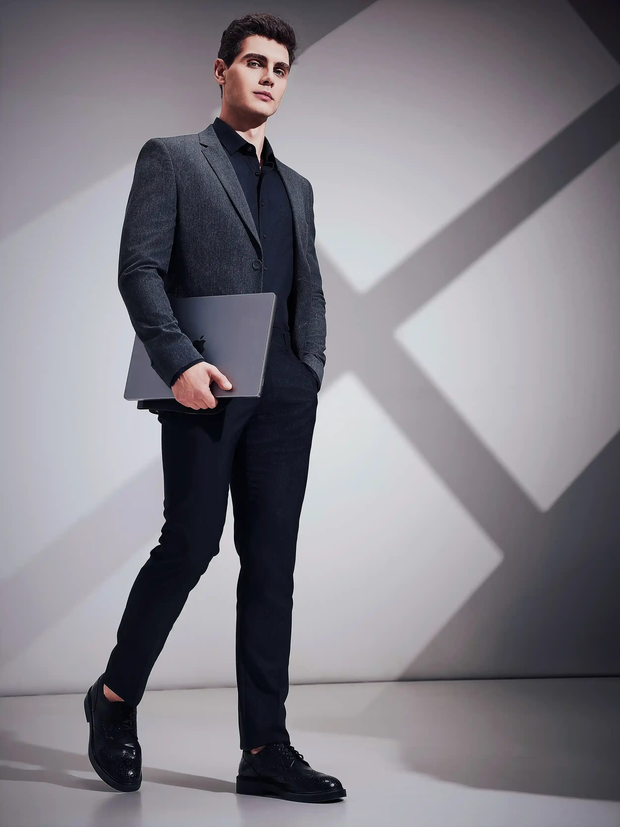 A young man with dark hair, dressed in a dark suit and black shoes, holding a closed silver MacBook in his right hand, standing in front of a modern abstract background. Anandclix, Bangalore, advertising photographer, Fashion photographer, Editorial.