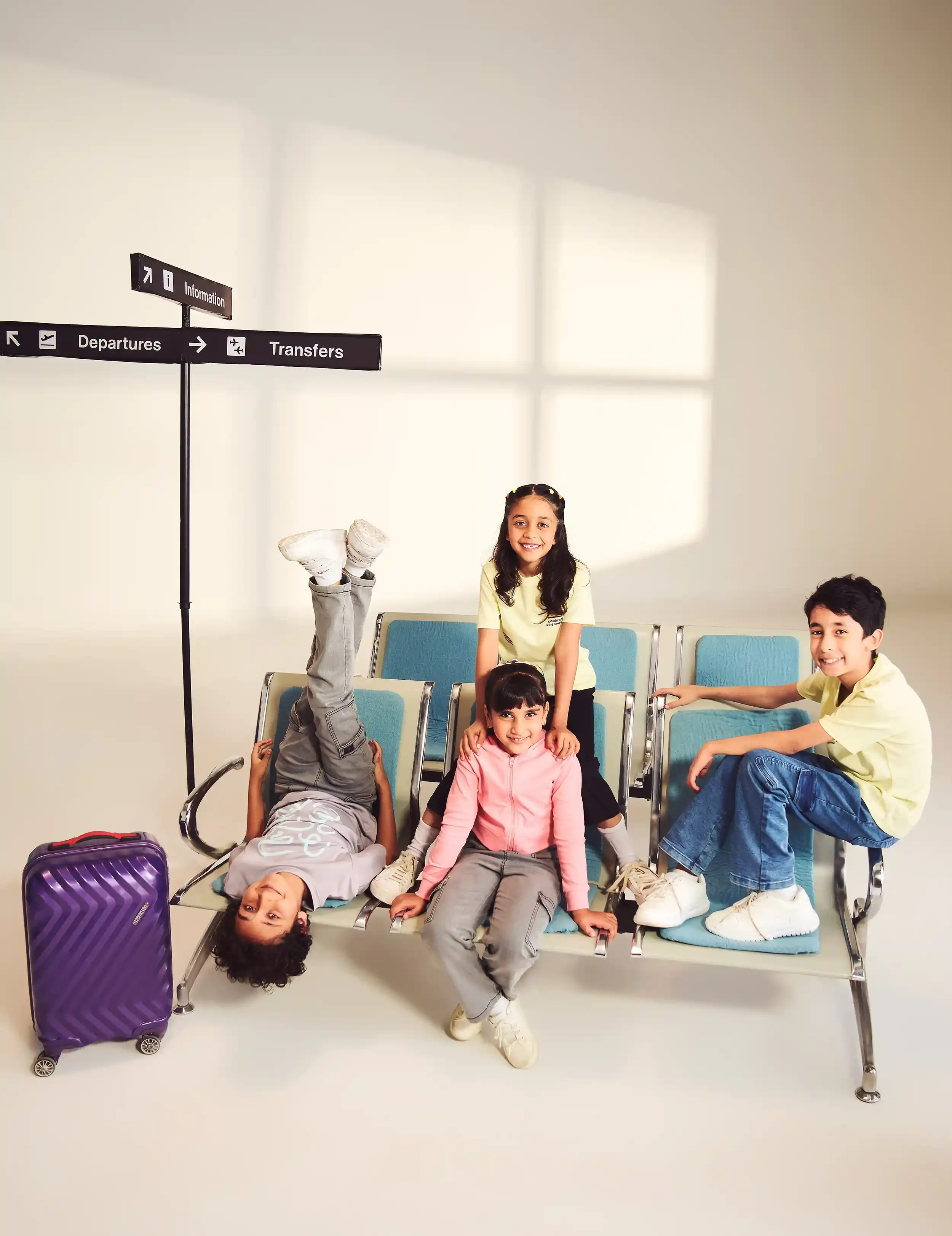 Four children sitting and lying on airport chairs with a purple suitcase nearby, in an airport waiting area with signage for departure, transfers, and information in the background.