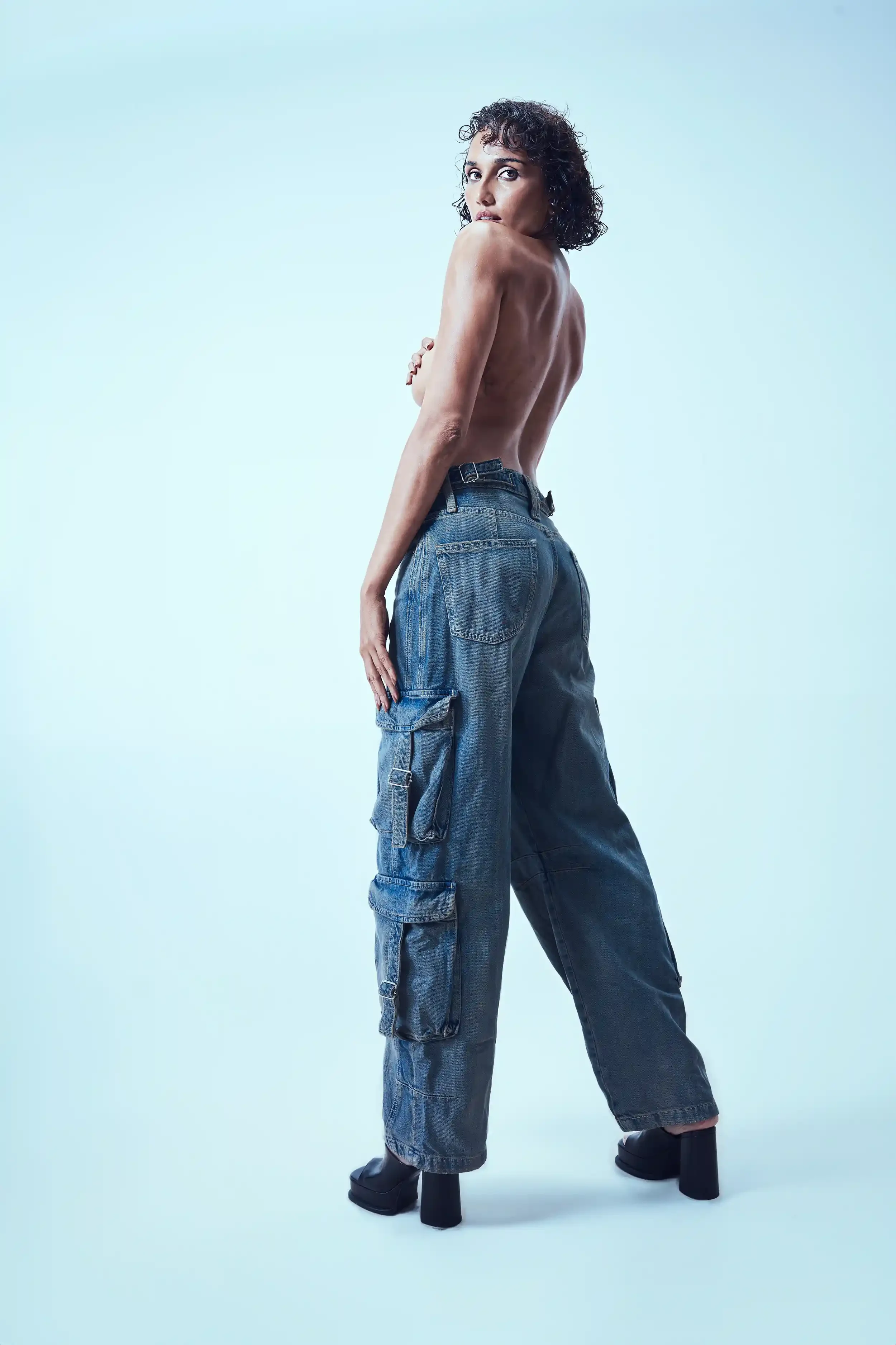A topless woman with short, curly hair poses wearing loose-fitting blue jeans and black high-heeled shoes against a plain light background.