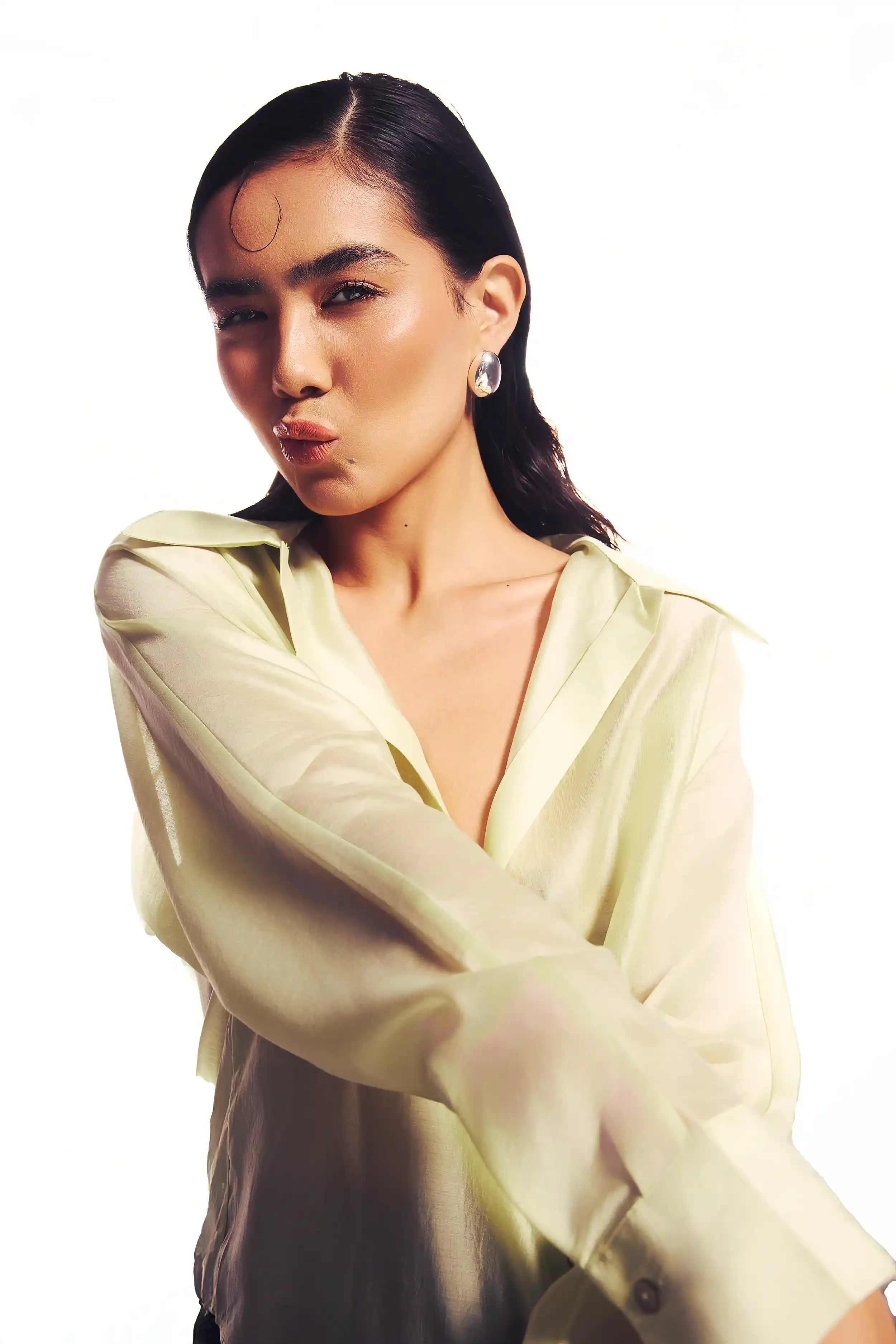 A woman with black, wet-look hair, wearing a shiny, pale yellow blouse, posing with her arms crossed, pouting, against a plain white background.