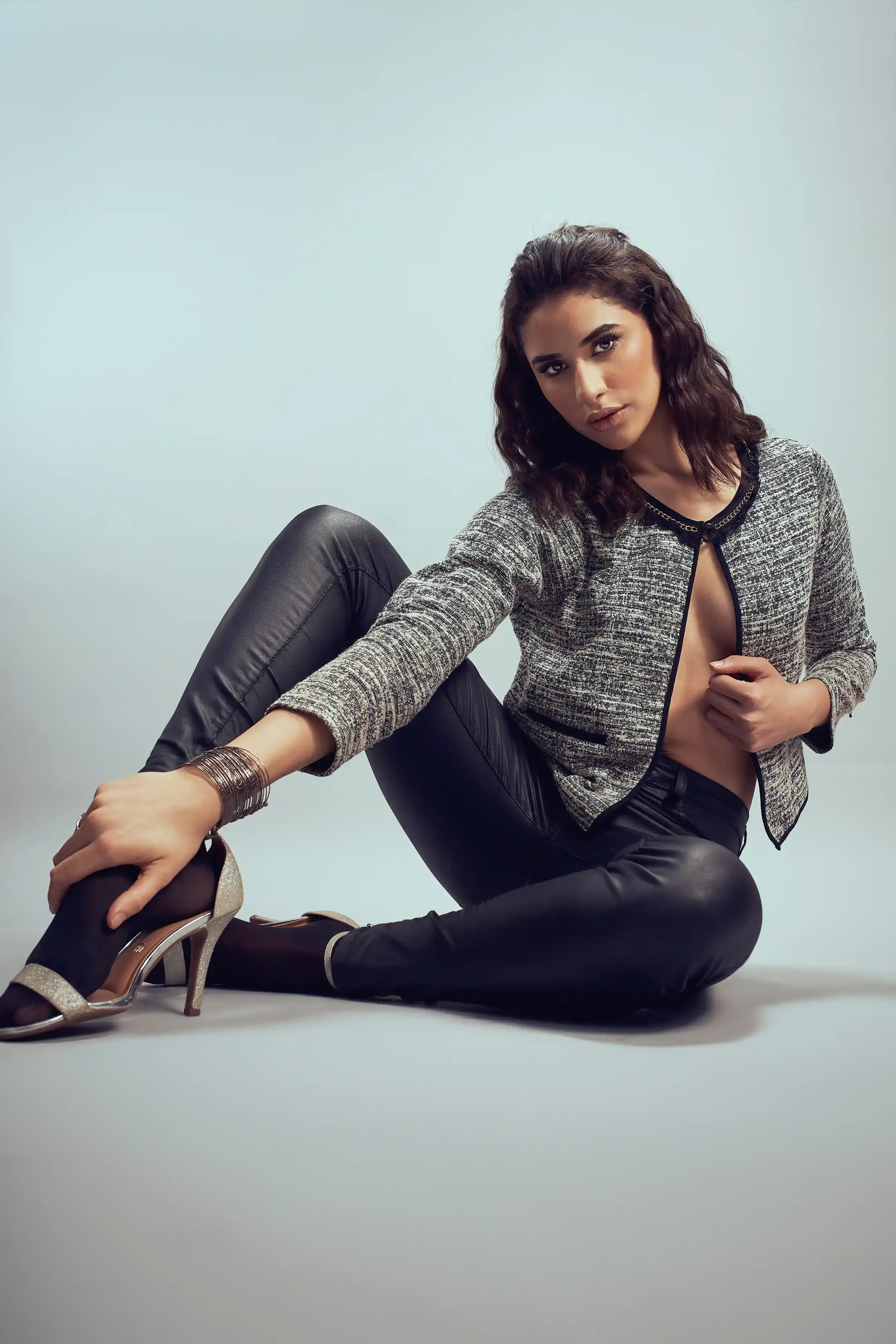 A woman with dark, wavy hair poses on the floor against a plain background, wearing a partially open blazer, black leather pants, and high heels.