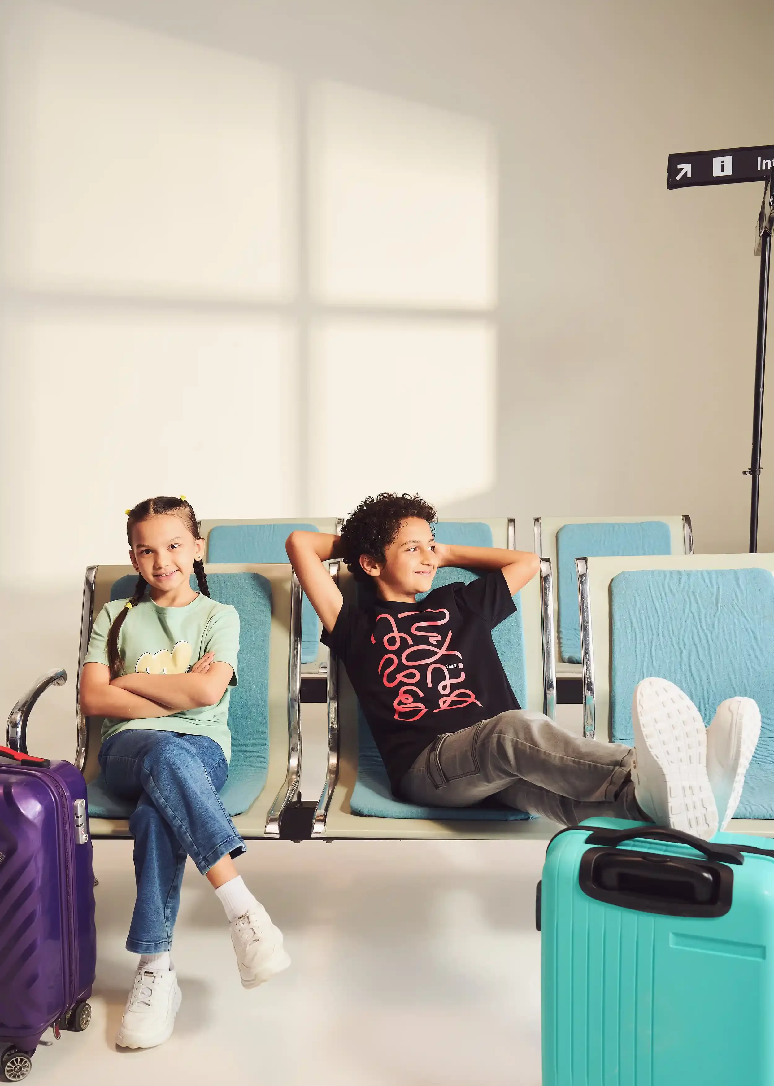 Two children sitting on airport chairs with luggage, a girl with braided hair in a green shirt, and a boy with curly hair in a black shirt, smiling, while waiting at an airport.