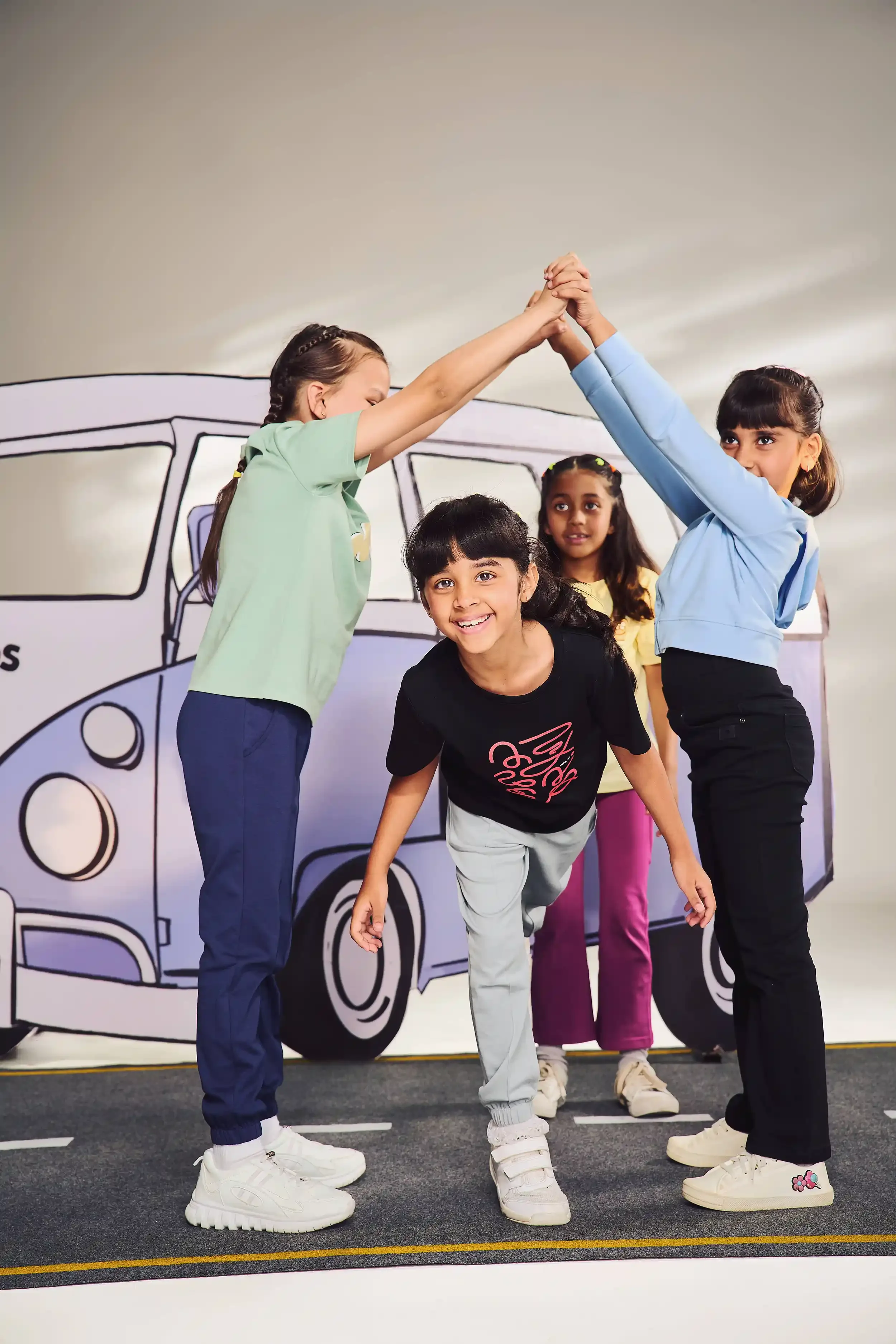 Four children with a cartoon bus backdrop, two girls high-fiving, one girl leaning forward, and one girl watching, all smiling.