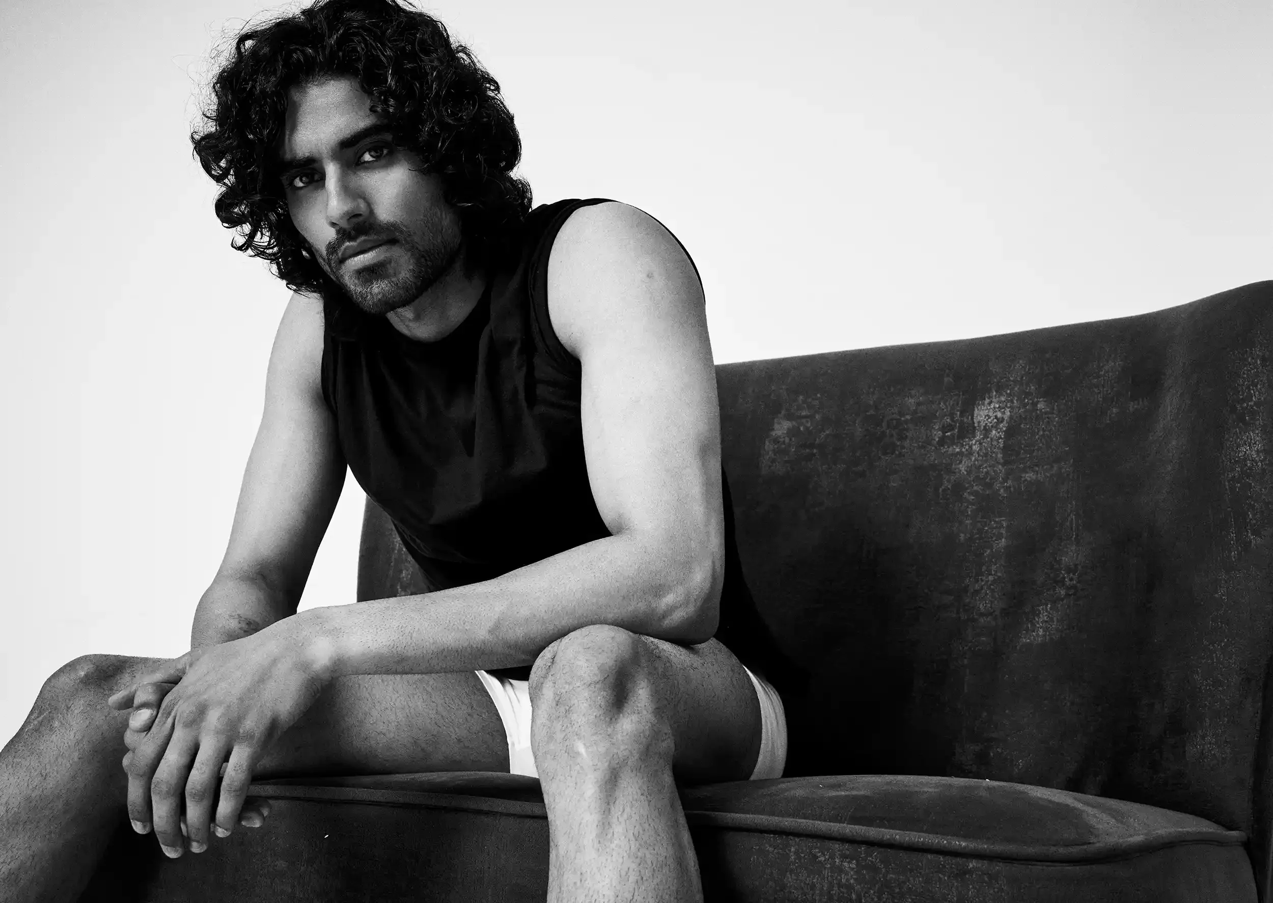 Black and white photo of a man with curly hair sitting on a dark couch, wearing a sleeveless shirt and shorts.