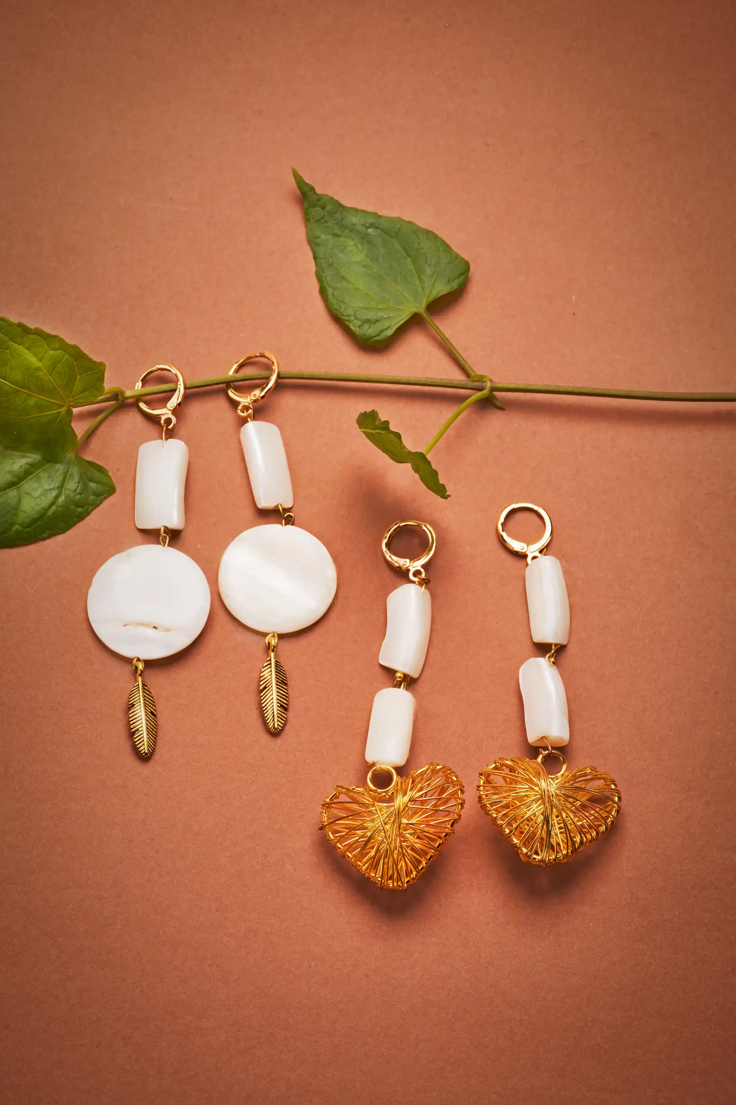 Two pairs of gold and white earrings with leaf and heart designs are displayed on a pink background with a green leaf.
