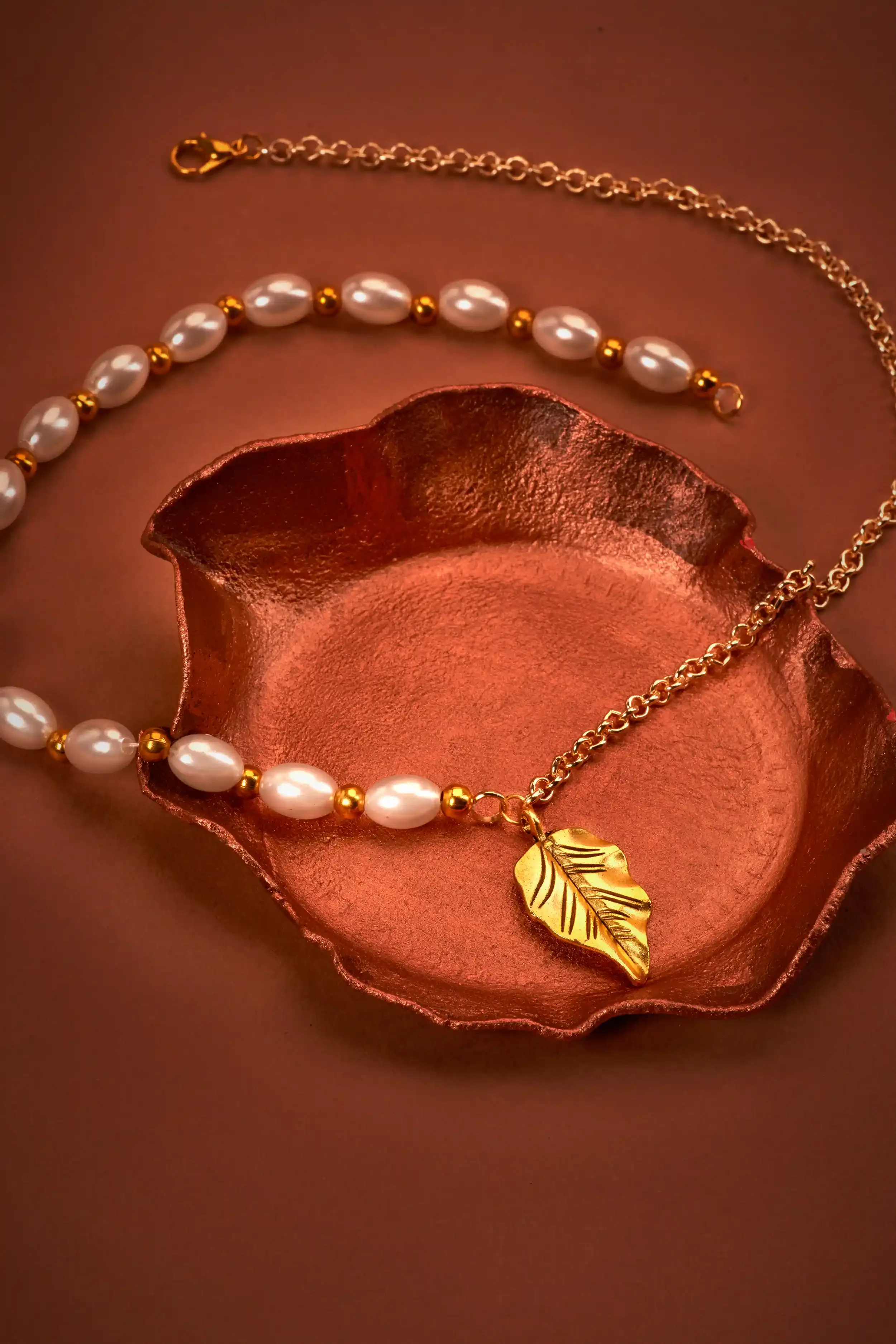 Gold necklace with a leaf pendant and a pearl necklace with gold beads placed in a copper bowl.