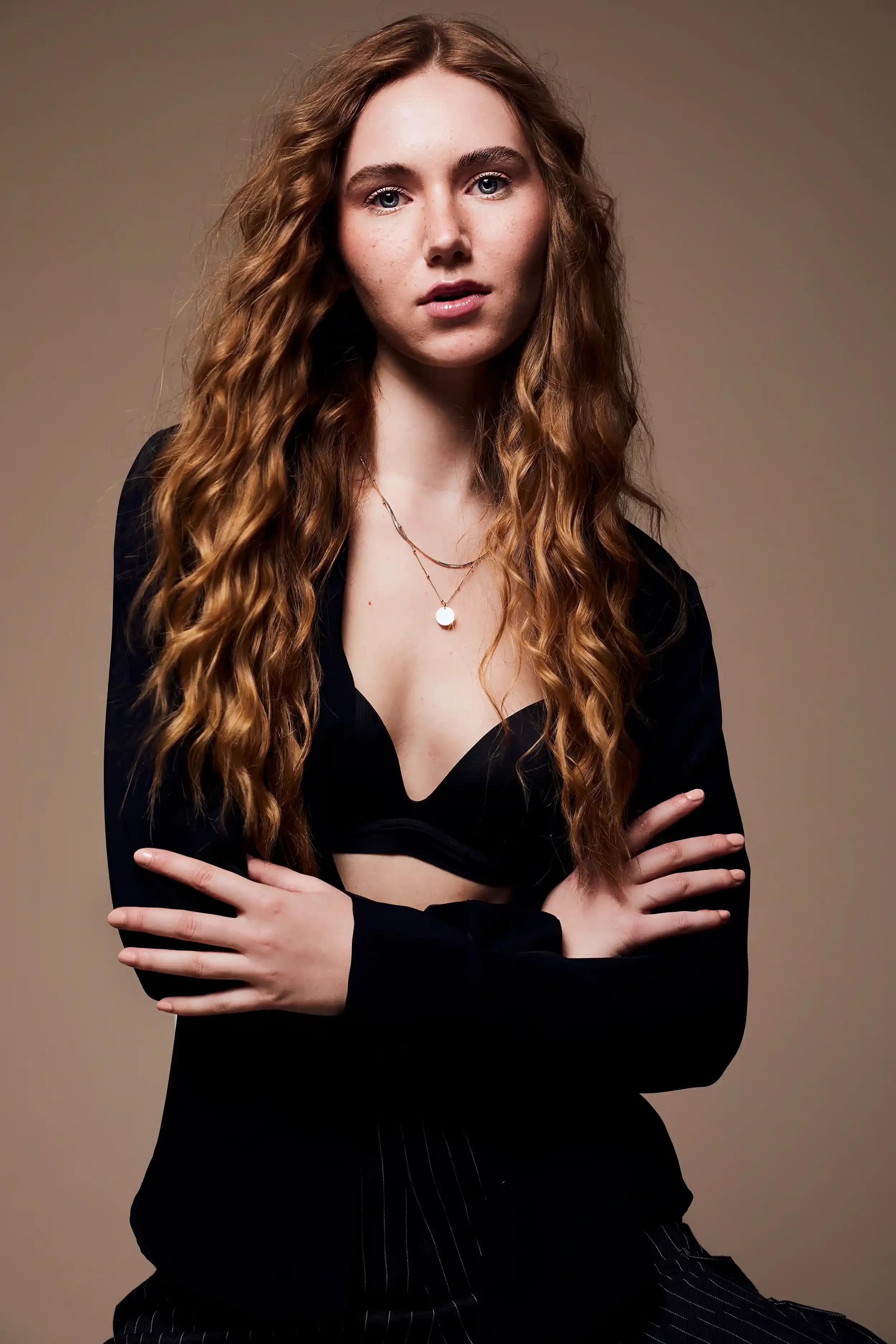 A woman with long, wavy red hair wearing a black top and layered necklaces, standing against a neutral background with arms crossed.