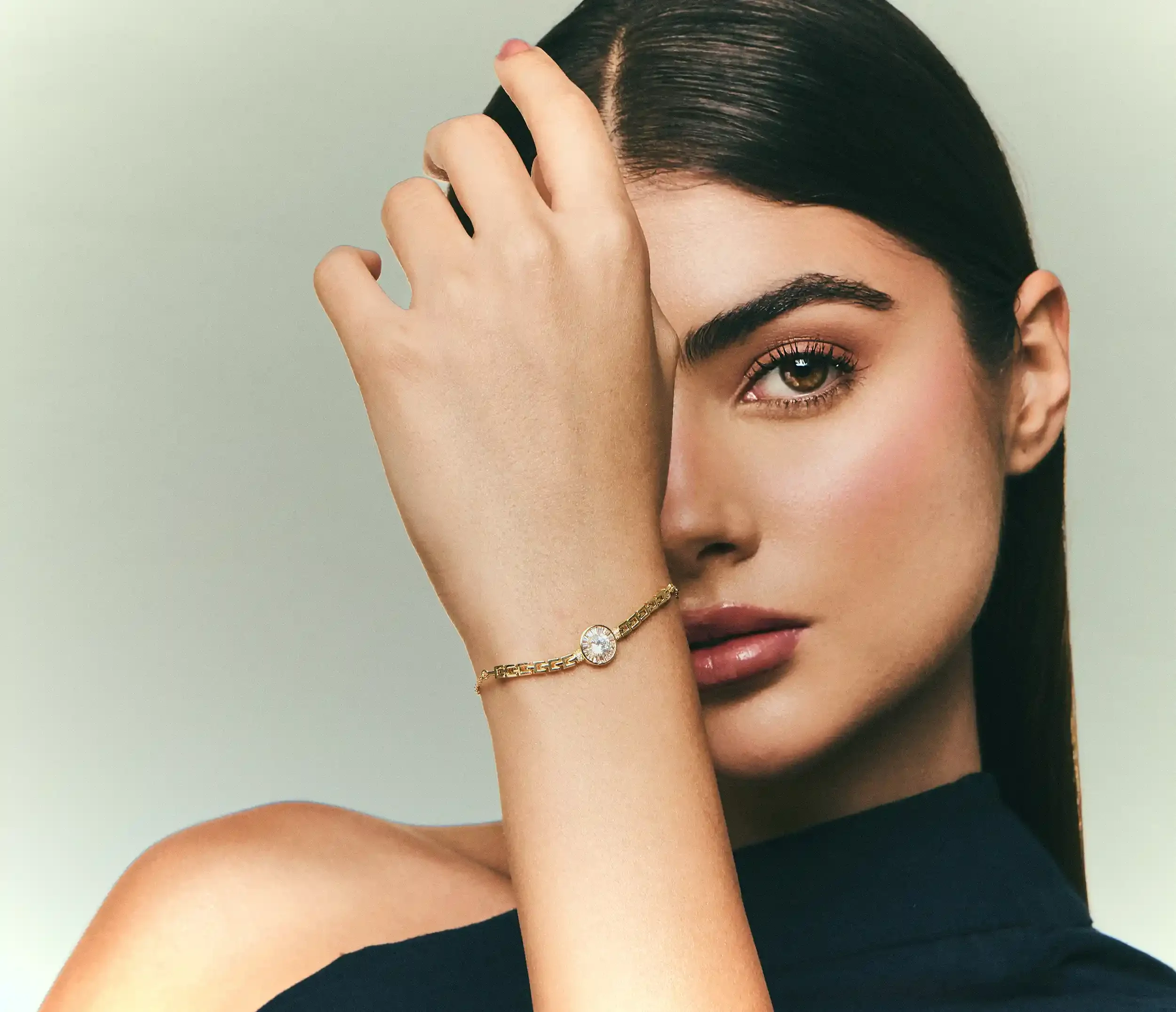 Close-up of a woman with long dark hair and brown eyes holding her hand over her face, showcasing a gold bracelet with a large clear gemstone. Kushal's Fashion Jewellery, Anandclix, Bangalore, advertising photographer, Fashion photographer.