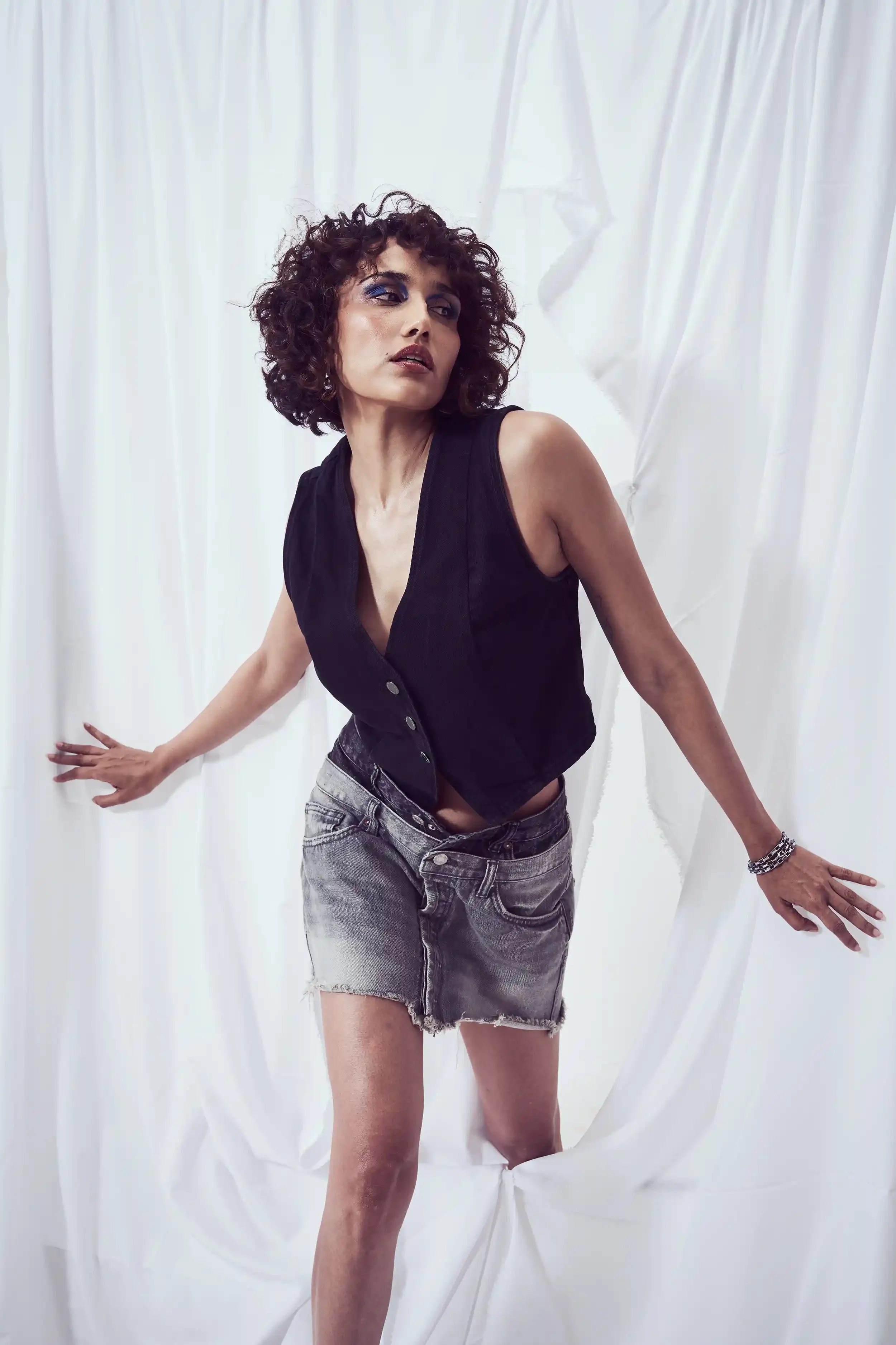 A woman with curly hair wearing a black sleeveless vest and frayed denim shorts standing against white draped fabric. Anandclix, Bangalore, advertising photographer, Fashion photographer, Editorial photographer, Bangalore photographer.