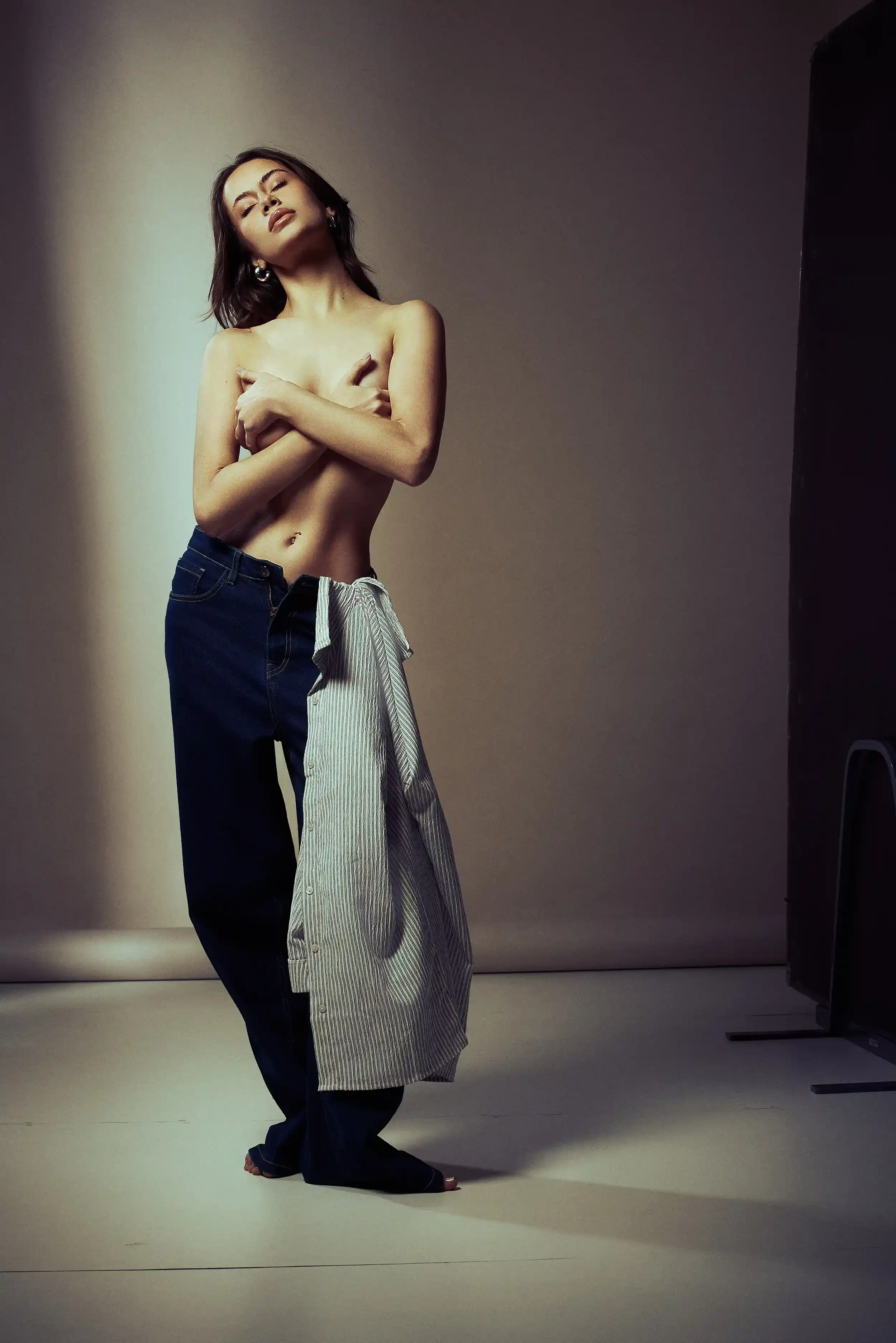 A topless woman with dark hair and earrings poses with her arms crossed in front of her chest, wearing loose jeans and a partially opened shirt hanging on her hips, in a studio setting.