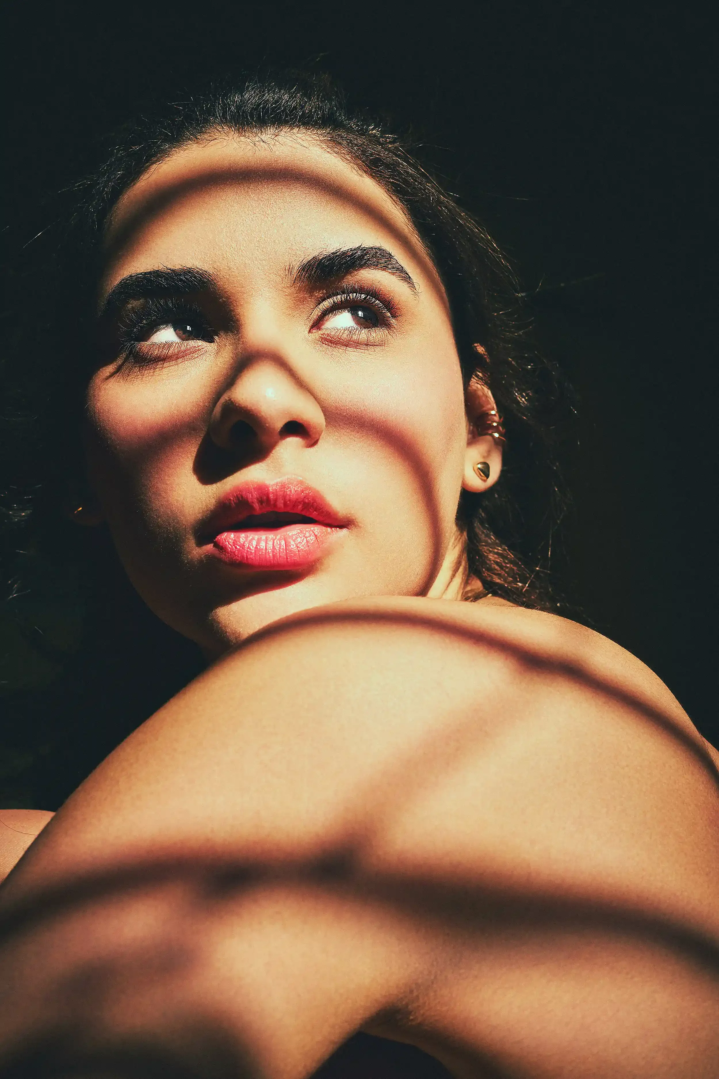 Close-up of a woman's face with dark hair, looking upward, illuminated by a focused light, with her shoulder and arm in the foreground. anandclix. Lingerie photography. Lingerie campaign. Fine Art Photography. Boudoir Photography.
