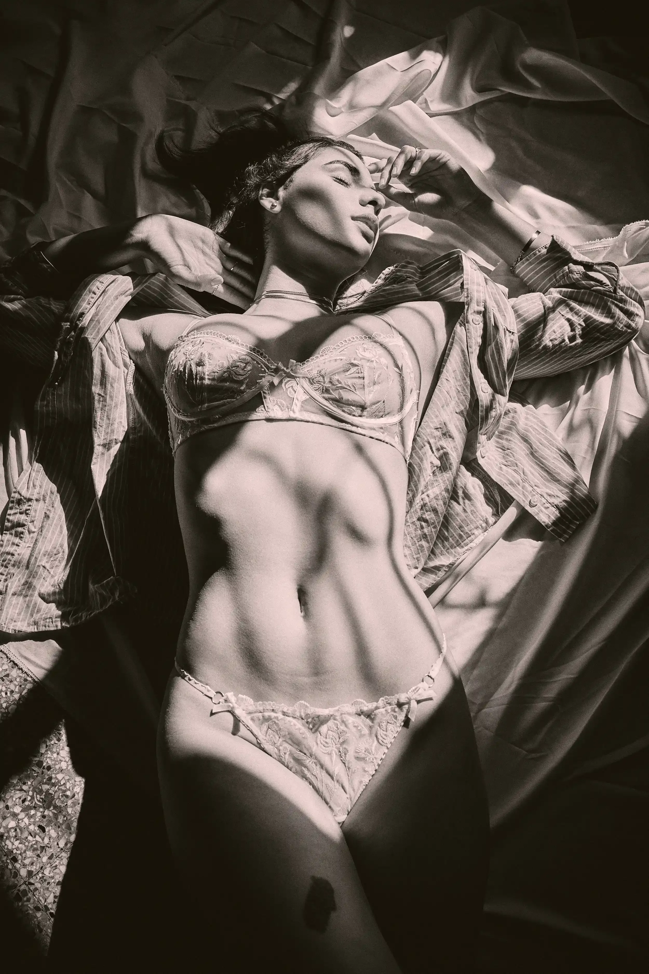 A woman with dark hair lying on her back on a bed with sheets and blankets. She is wearing lace lingerie and a striped shirt. She has her eyes closed, with one hand near her face and the other resting on her head. anandclix. Lingerie photography. 