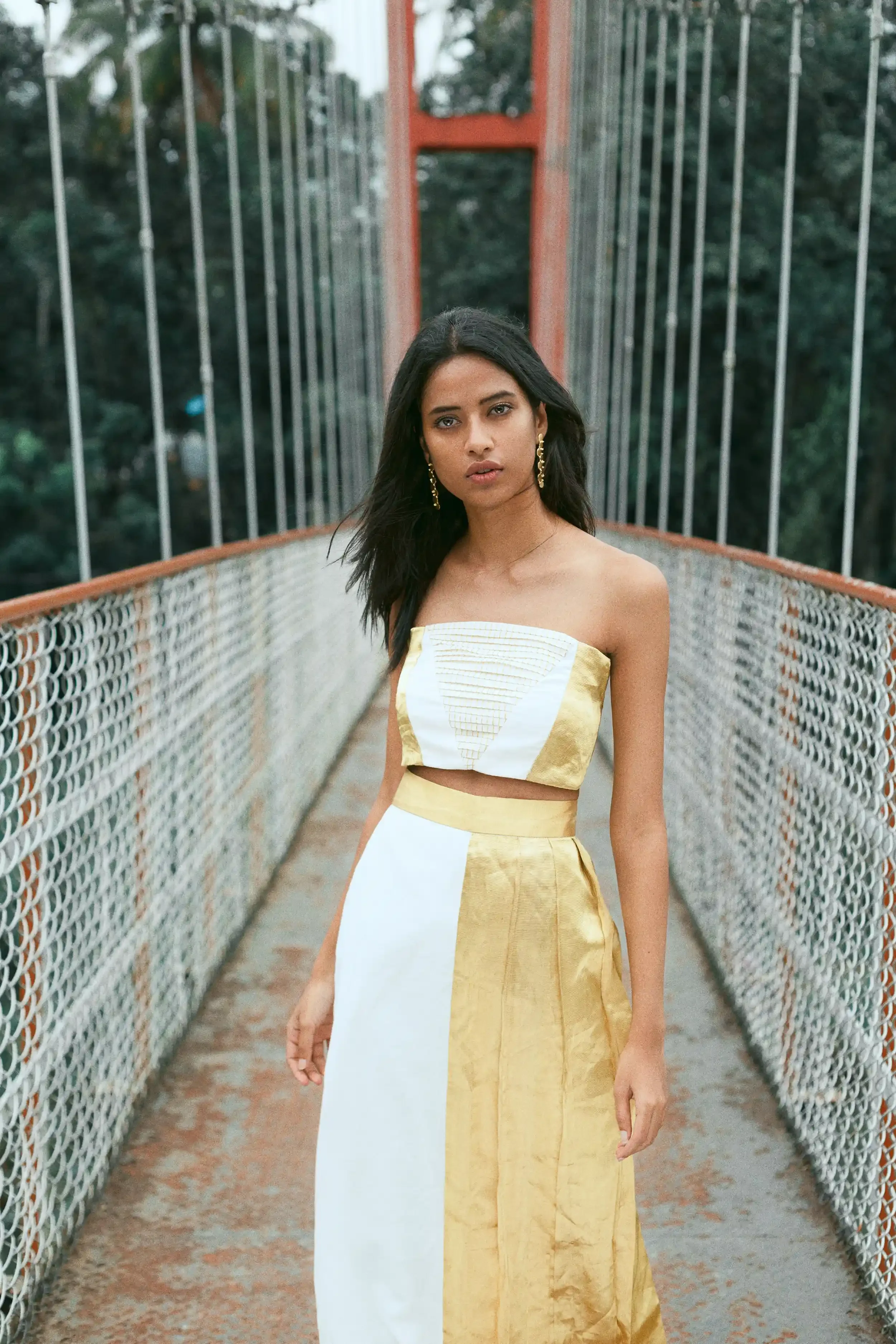 A young woman with dark hair and earrings standing on a bridge with tall railings and a red support structure.