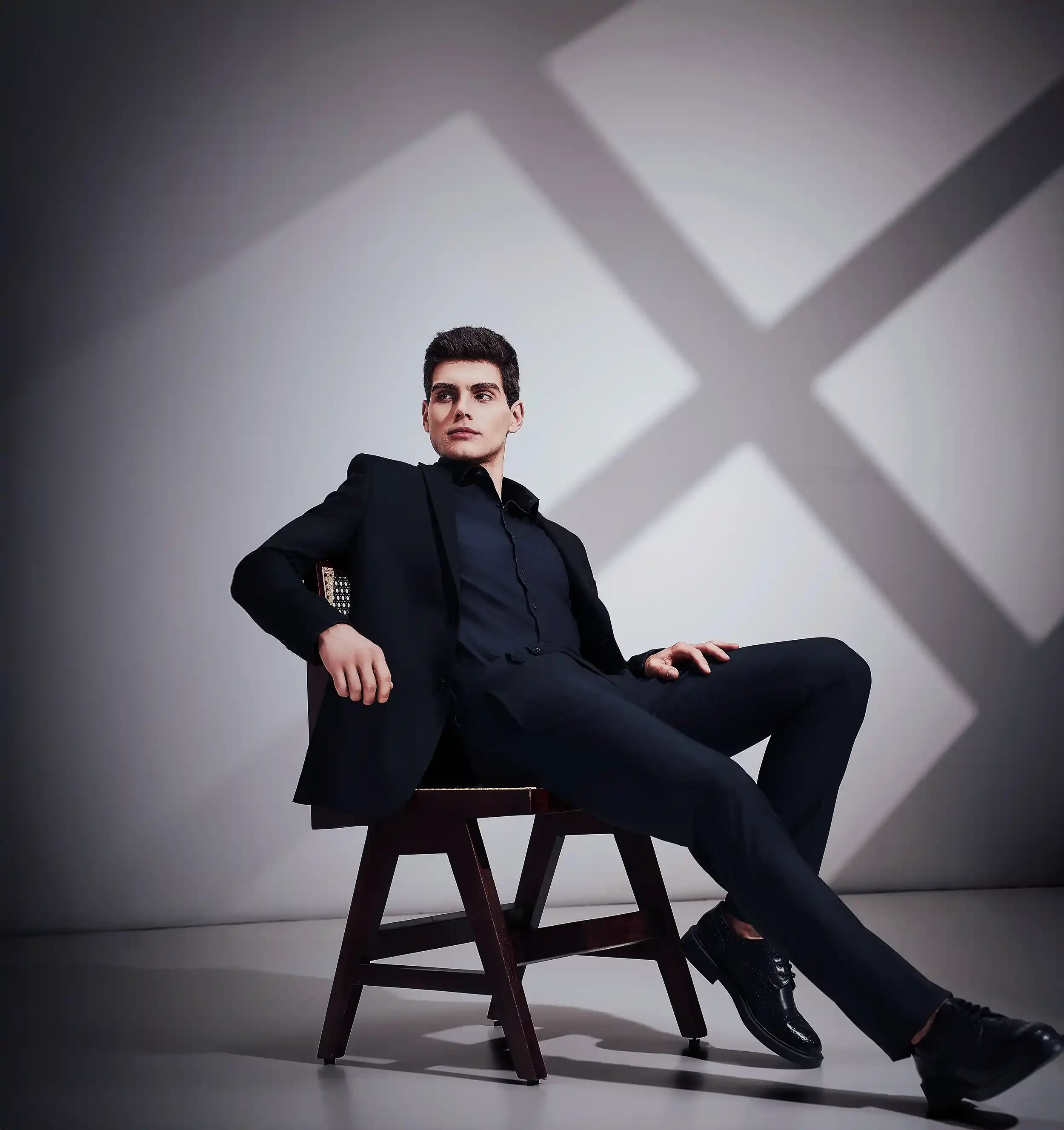 A young man in a black suit and dark shirt sitting on a wooden chair with one arm resting on the chair's armrest and one hand on his thigh, in a studio with a light background and shadow patterns cast on the wall behind him.