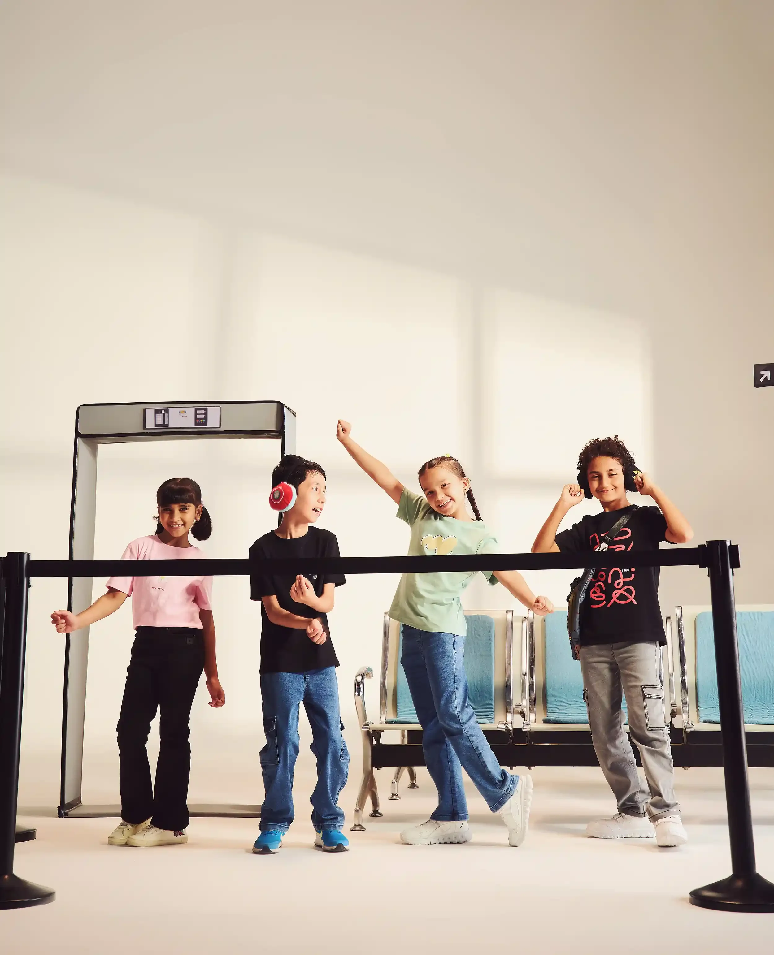 Four children at an airport, waiting behind a queue barrier, having fun and listening to music with headphones.