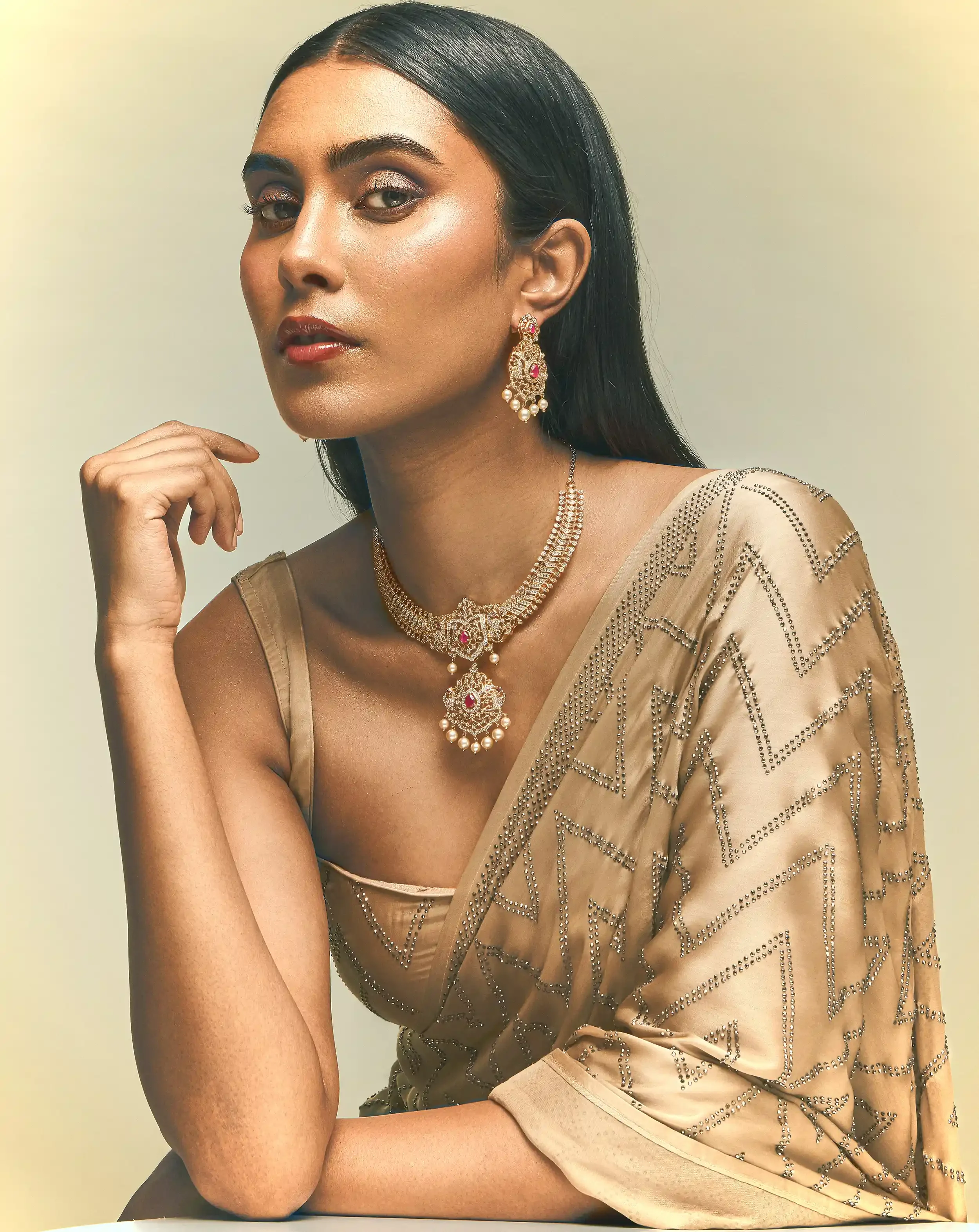 A woman with long black hair styled straight and sleek, wearing glittery makeup, red lipstick, and ornate gold jewelry including earrings, necklace, and bracelet, dressed in a beige sari with silver embellishments, posing with her chin resting on her hand against a neutral background.