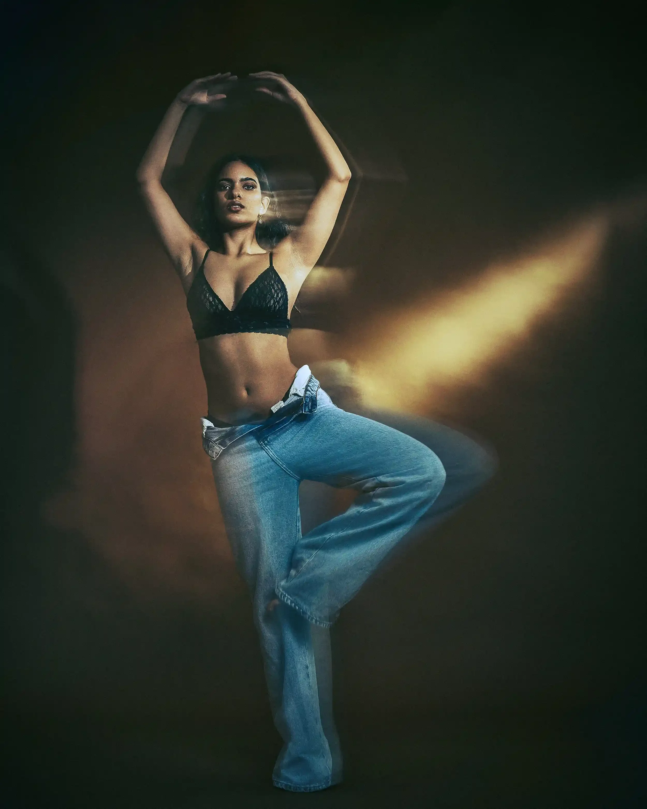 A woman dancing in a dark, smoky setting, wearing a black lace crop top and loose blue jeans, with her arms raised above her head.