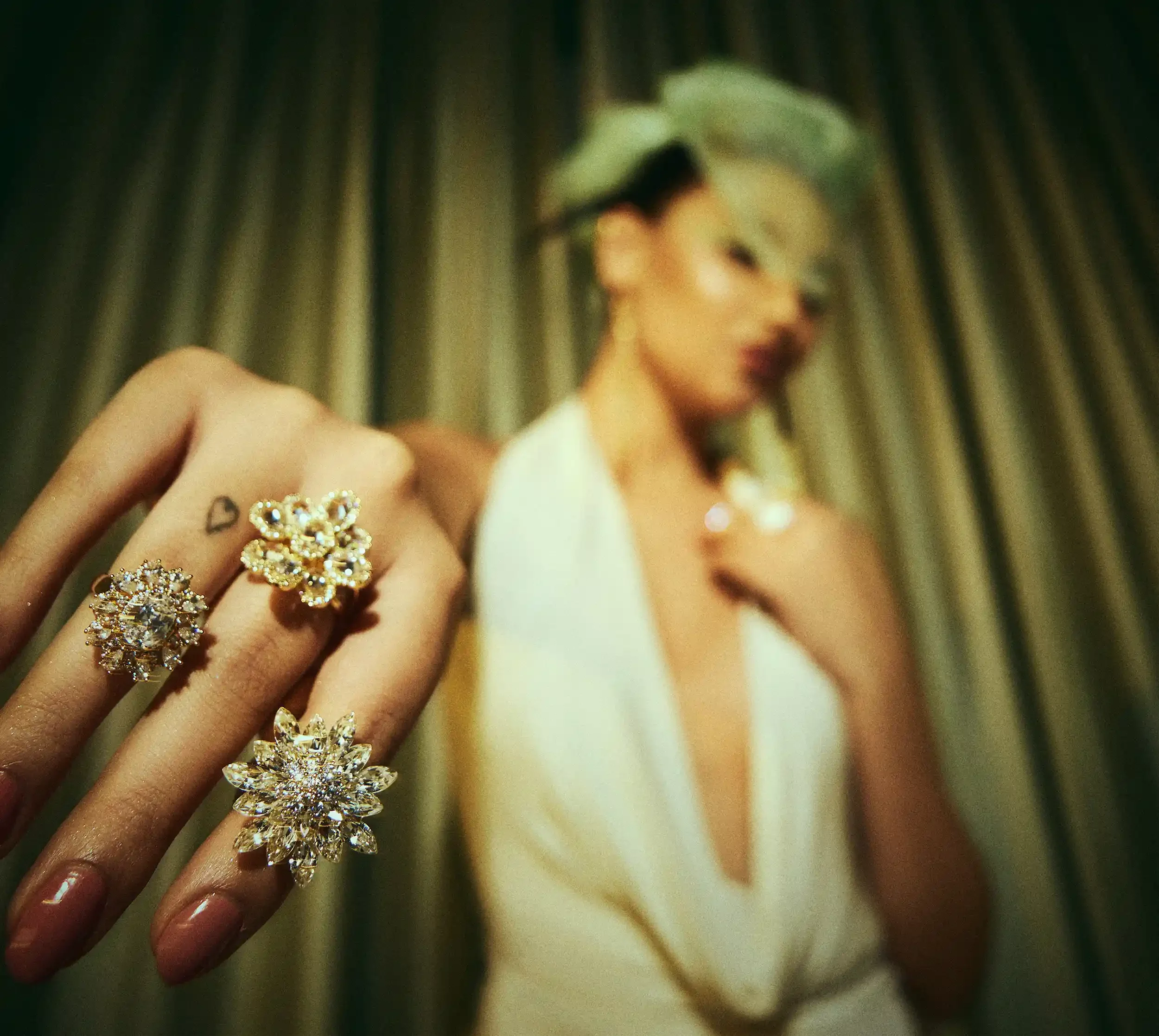 Close-up of four sparkling rings with floral and gem designs on a woman's fingers, with a woman in a white dress with a deep neckline and blonde hair in the background.