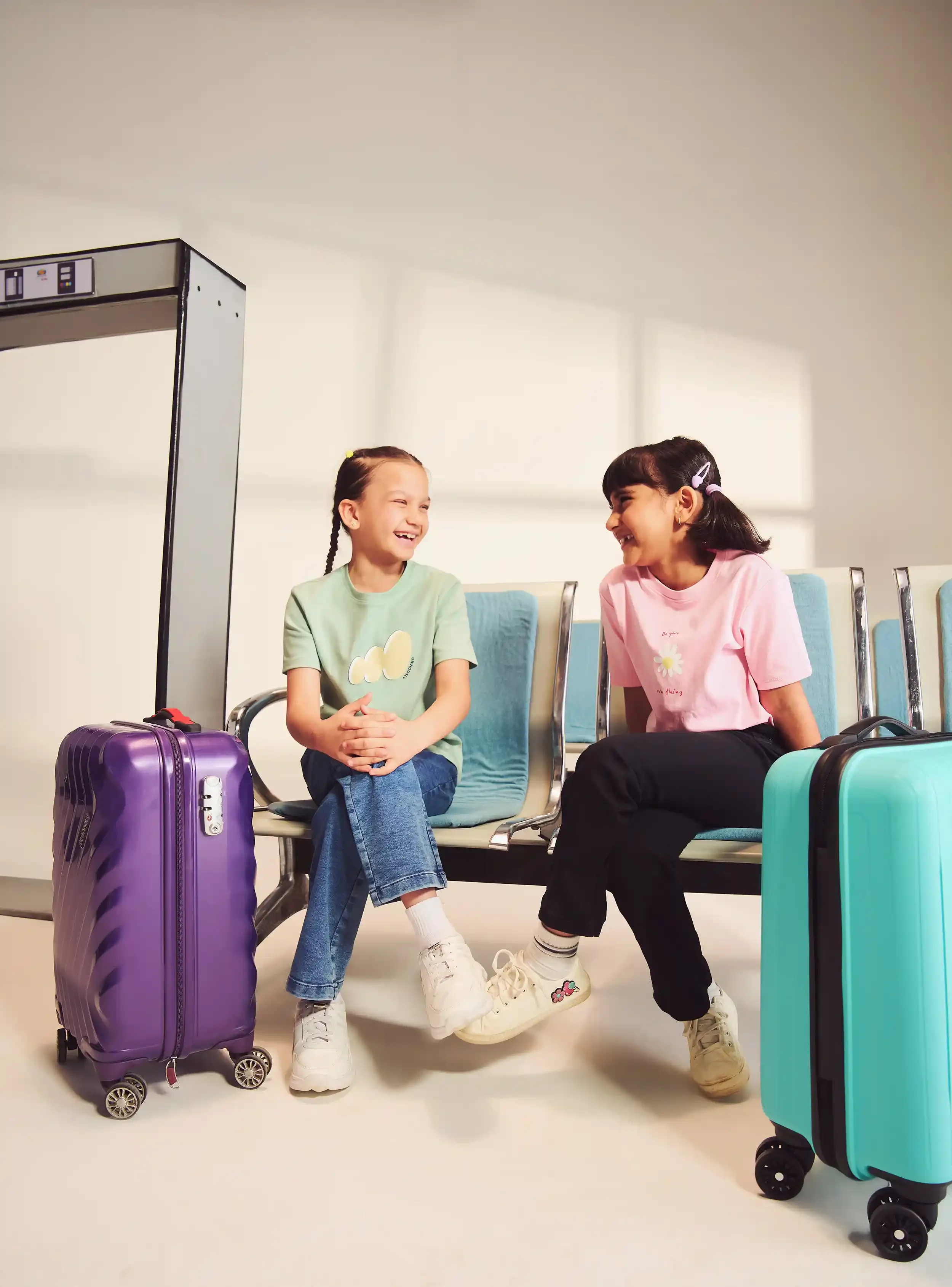 Two young girls sitting on airport chairs, smiling and laughing at each other, with colorful suitcases in front of them.