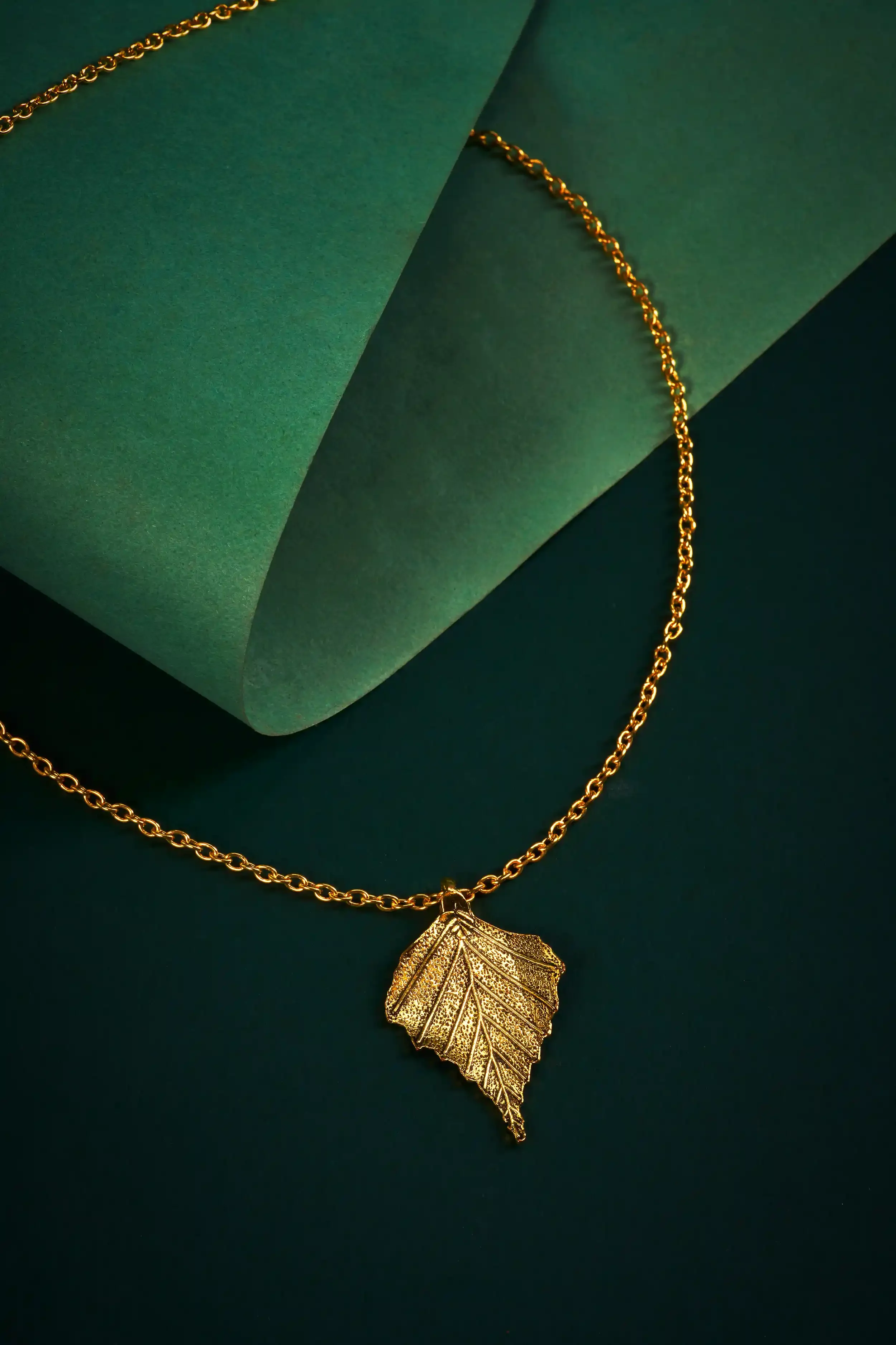 A gold necklace with a leaf-shaped pendant resting on a dark green surface, partially leaning against a rolled green paper.